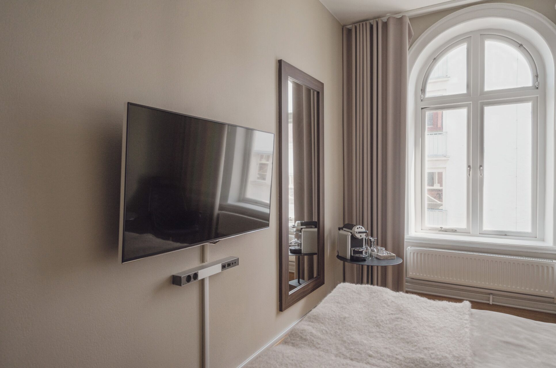 Habitación de hotel con televisor de pantalla plana en la pared, un espejo grande, un radiador y una ventana con cortinas.