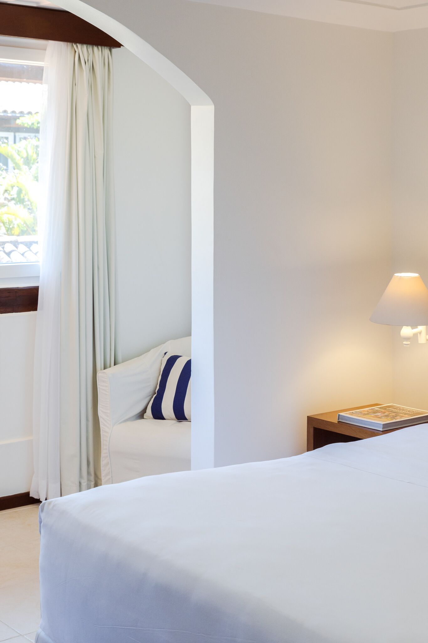 A hotel room with a bed, a nightstand with a lamp, a window with curtains, and a view of greenery outside.