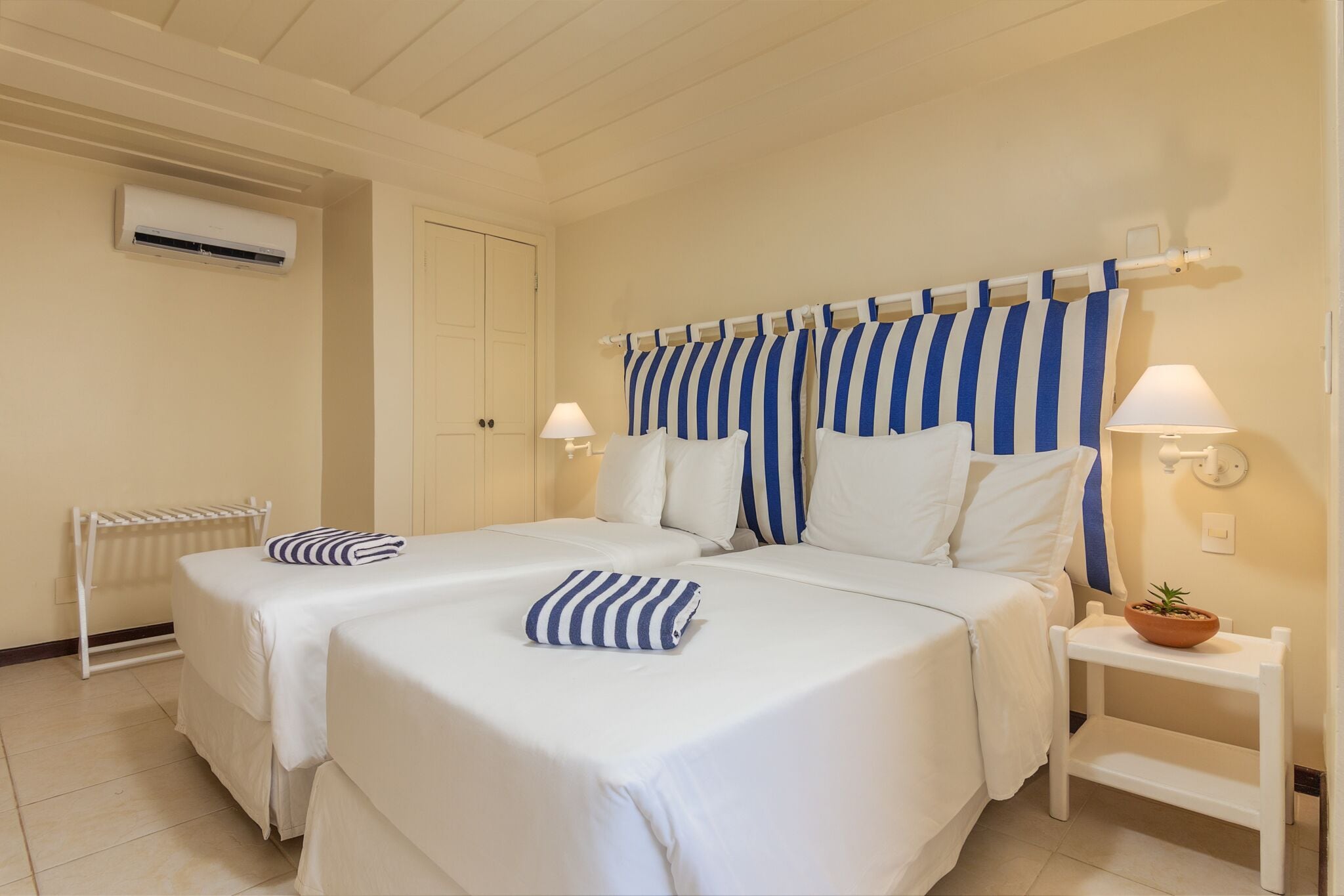Habitación de hotel con dos camas, ropa de cama blanca, almohadas a rayas, una mesita con lámpara y aire acondicionado instalado en la pared.