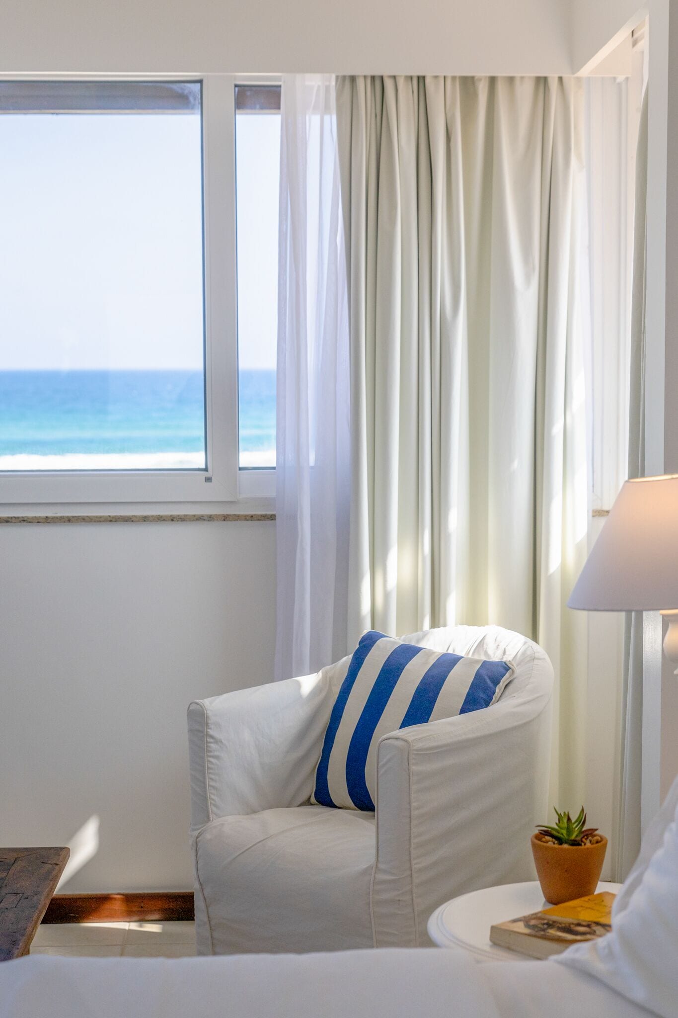 A room with a view of the ocean, a white chair with a blue and white striped pillow, a small potted plant, and a lamp on a side table.