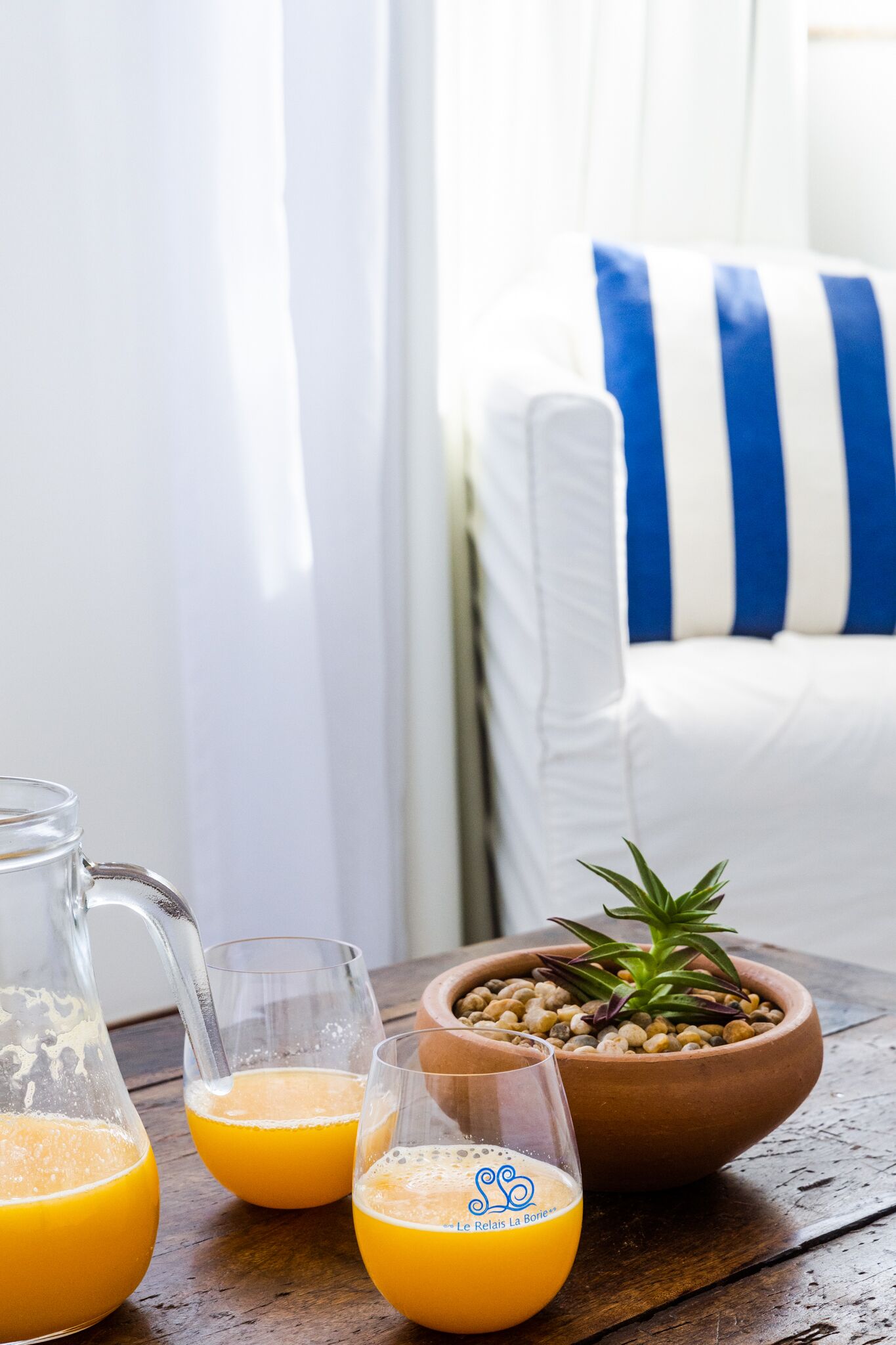 Un desayuno servido con jugo de naranja en vasos y una jarra, un tazón de nueces y una pequeña planta sobre una mesa de madera.