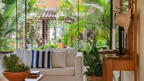 A cozy living room with a white sofa, a large potted plant, and a view of a tiled roof through a window. - Le Relais La Borie (Búzios)