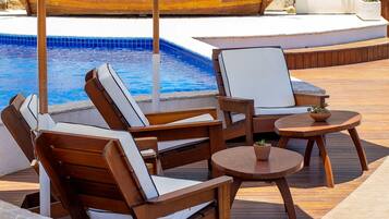 A poolside area with wooden lounge chairs and a table, a stone wall, and a boat in the background.