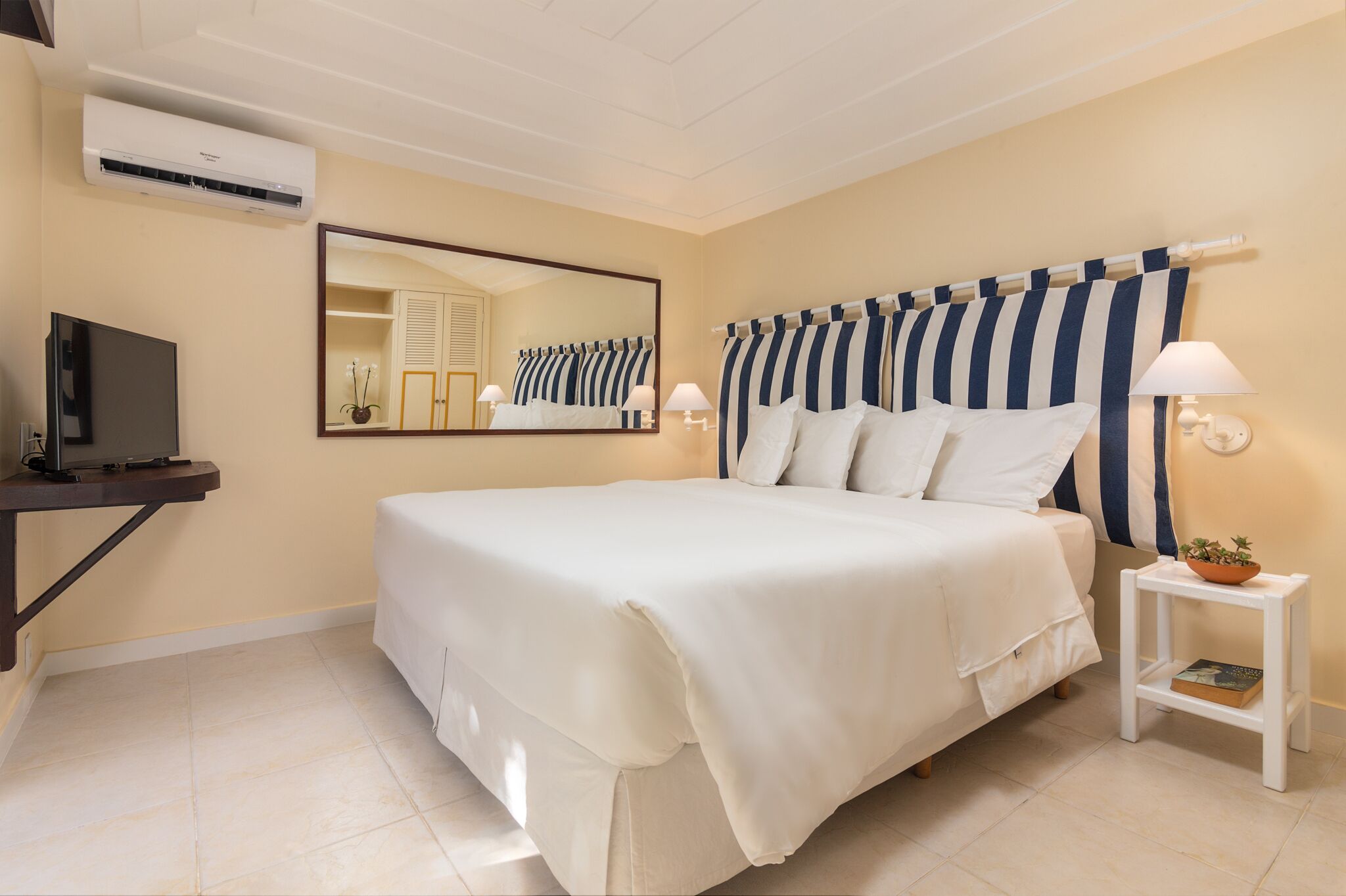 A hotel room with a large bed, a TV on a stand, a mirror, a bedside table with a lamp, and a small plant.