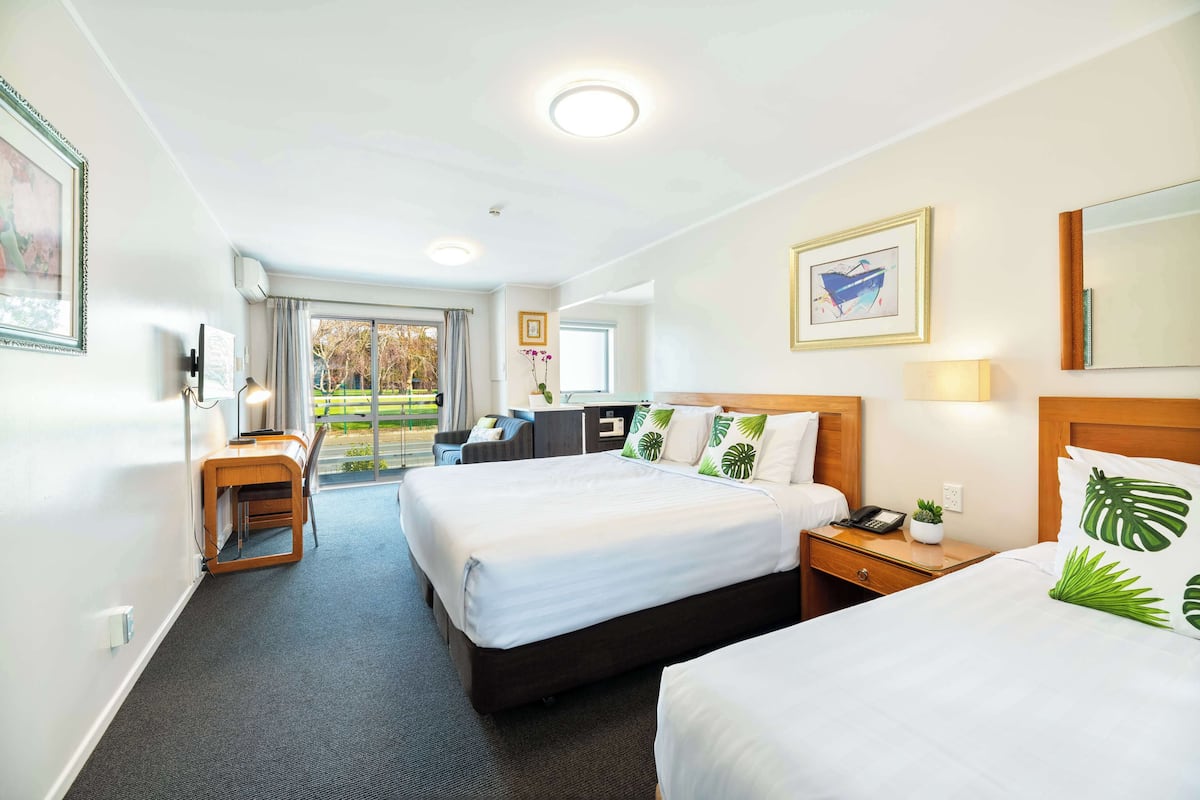 A hotel room with two beds, a desk, a chair, a small table with a lamp, and a view of the outdoors.
