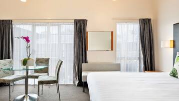 Habitación de hotel con cama, una mesita de vidrio con sillas, una planta en maceta y vistas de la ciudad a través de amplios ventanales.