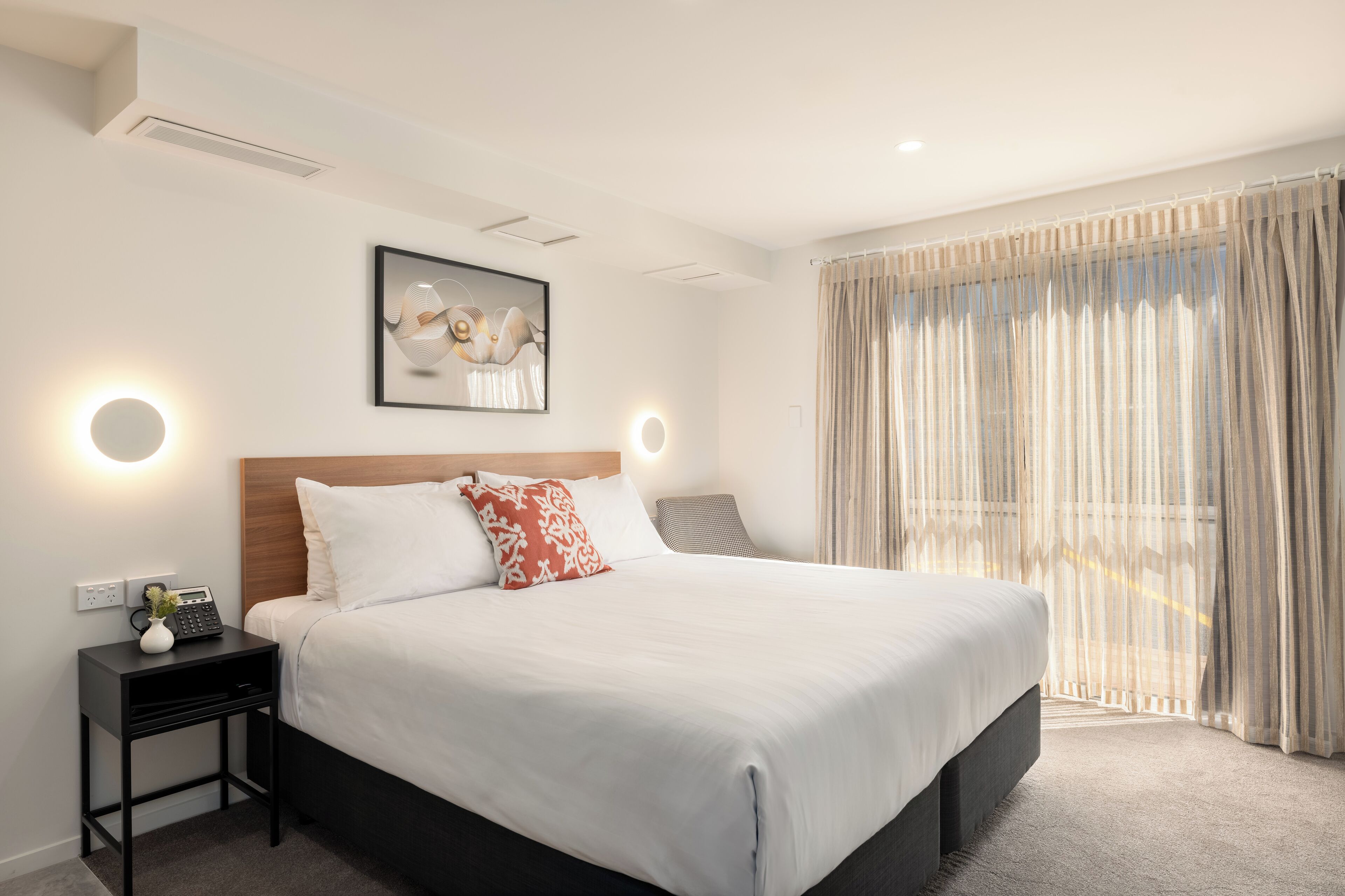 A neatly made bed with white linens and a red pillow, a bedside table with a lamp, and a framed artwork above the bed.