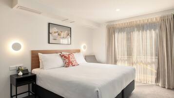 A neatly made bed with white linens and a red pillow, a bedside table with a lamp, and a framed artwork above the bed.
