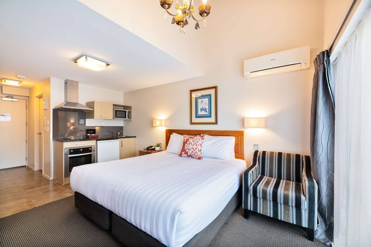 A hotel room with a large bed, a striped armchair, a kitchenette, and a ceiling light fixture.