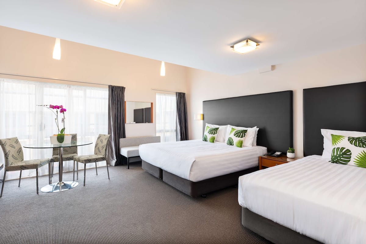 A hotel room with two beds, a sitting area with a glass table and chairs, and a large window with curtains.