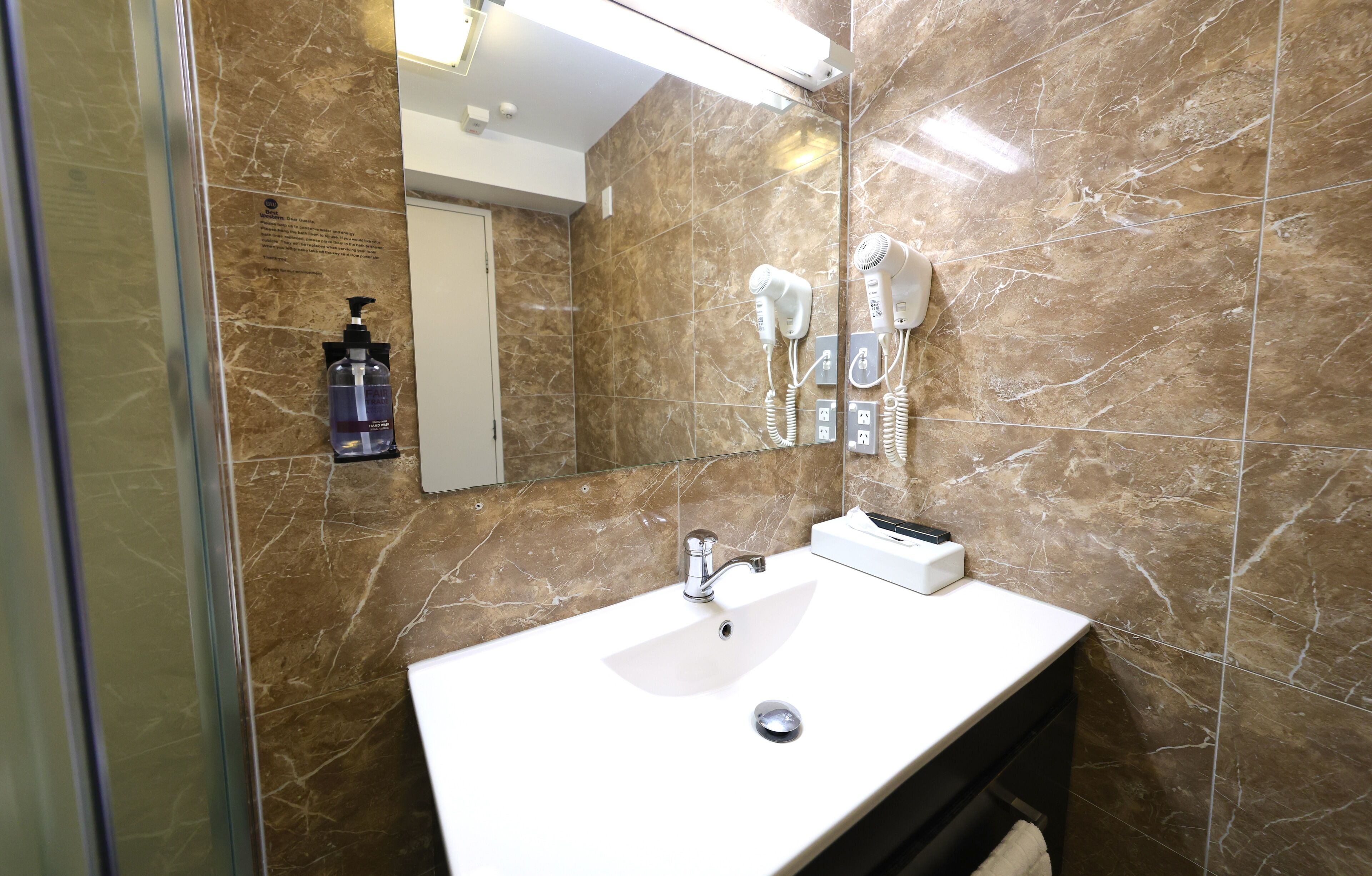 A bathroom with a marble wall, a sink, a mirror, a hairdryer, and a bottle of hand soap.
