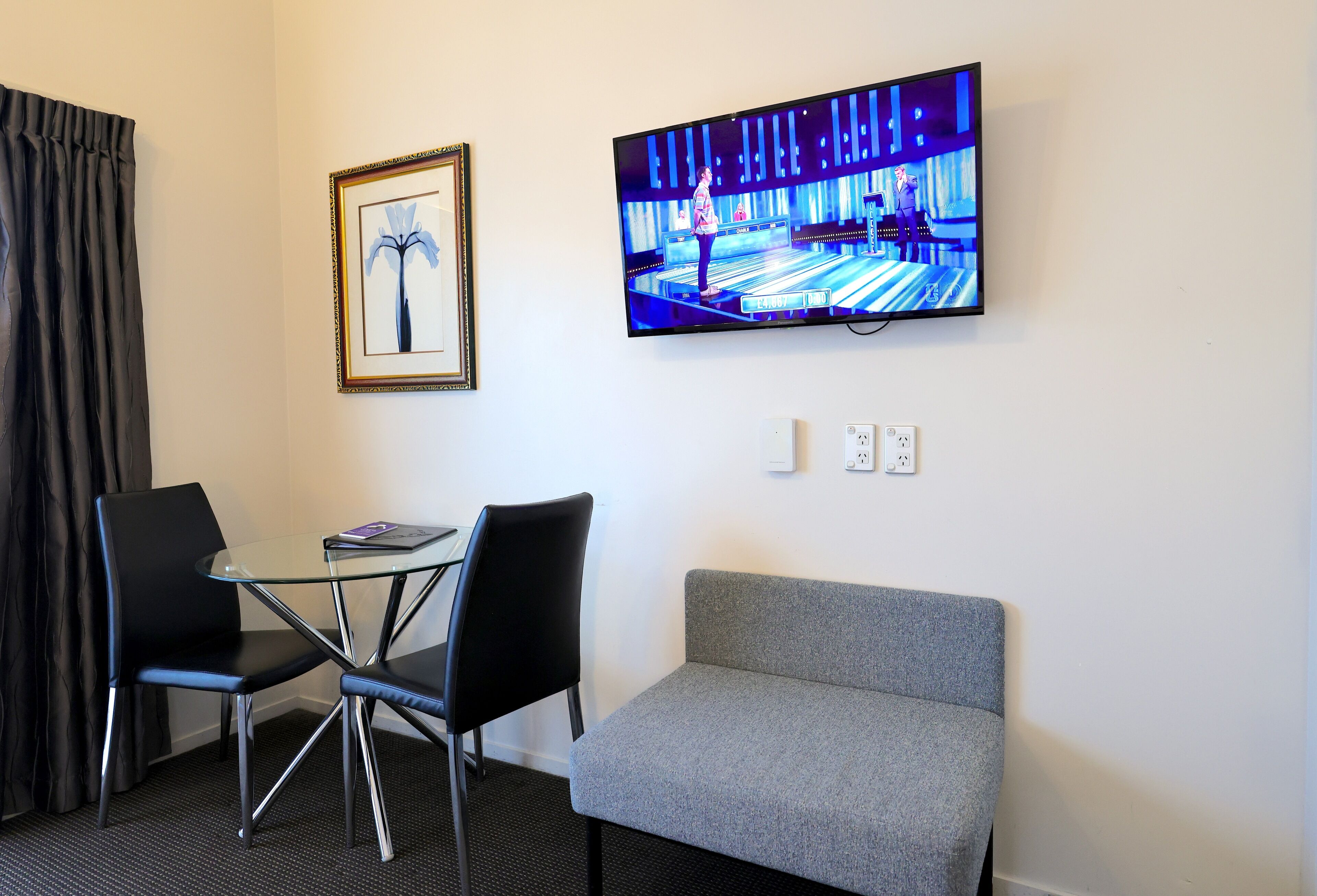A hotel room with a television displaying a bowling game, a framed picture on the wall, a grey sofa, and two black chairs around a small glass table.
