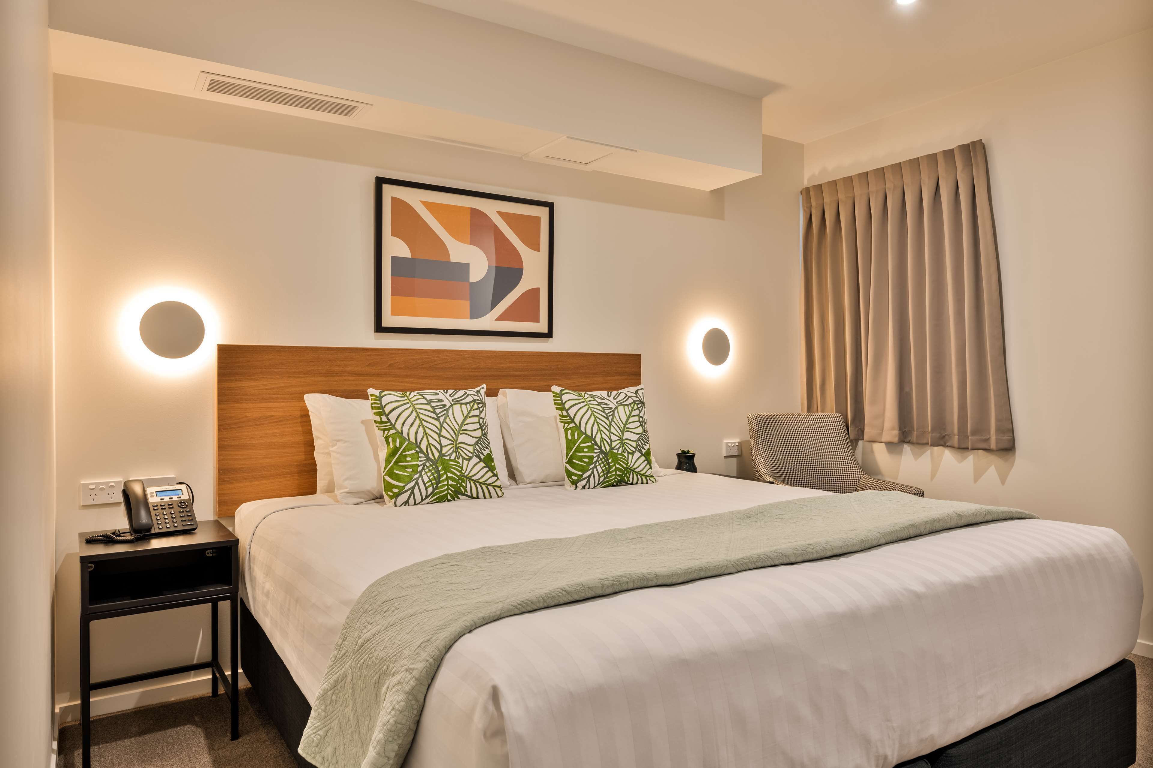 A hotel room with a large bed, a bedside table with a phone, a headboard with a leaf-patterned pillow, a wall-mounted abstract art piece, and a window with curtains.
