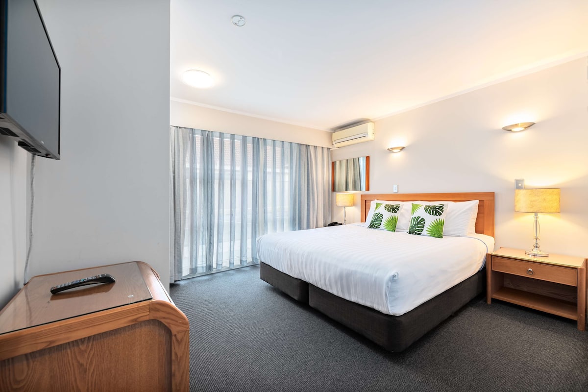 A hotel room with a bed, bedside tables, a TV, and a window with curtains.
