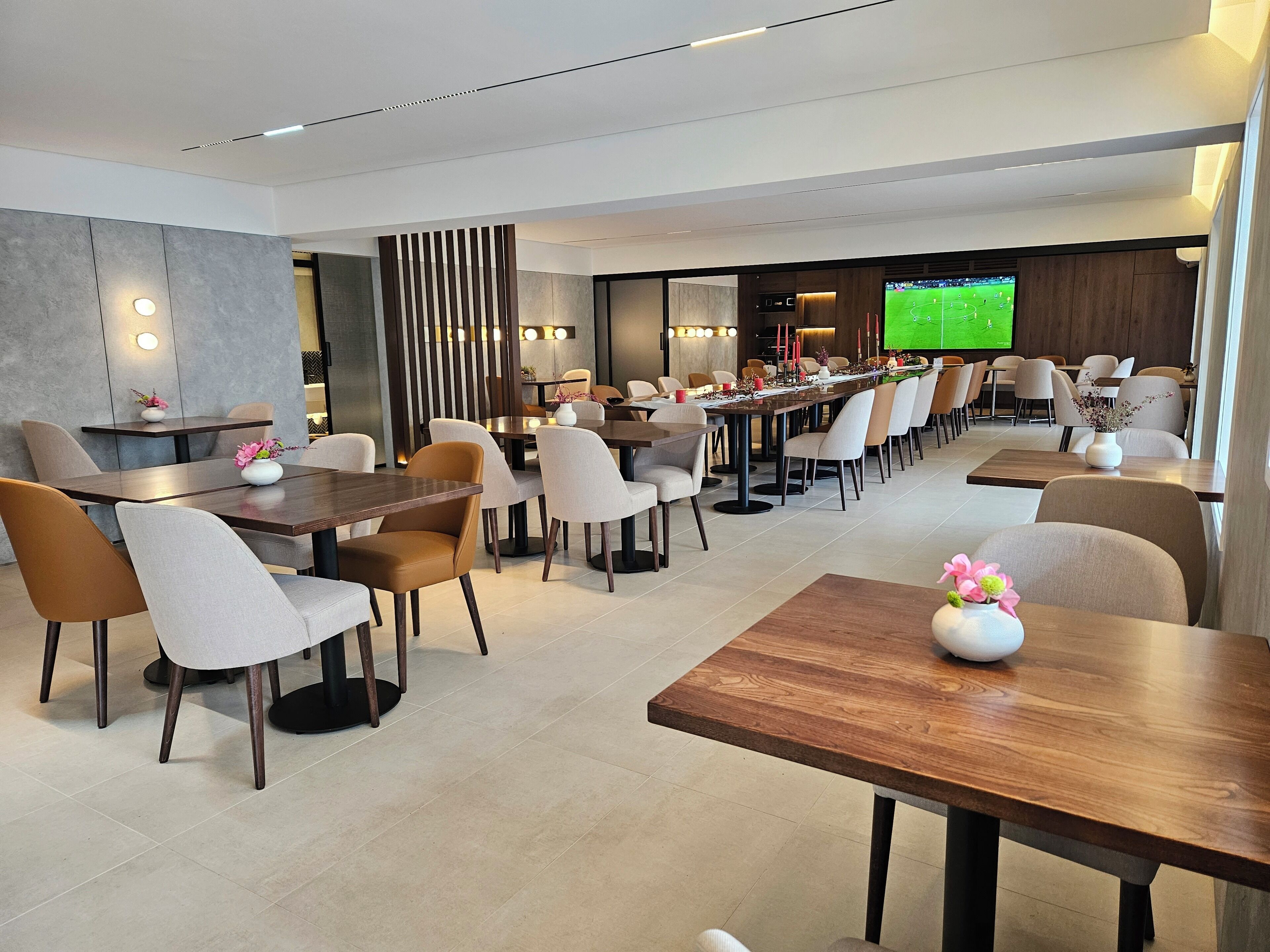 A modern dining area with multiple tables and chairs, a long communal table, and a television screen displaying a sports game.