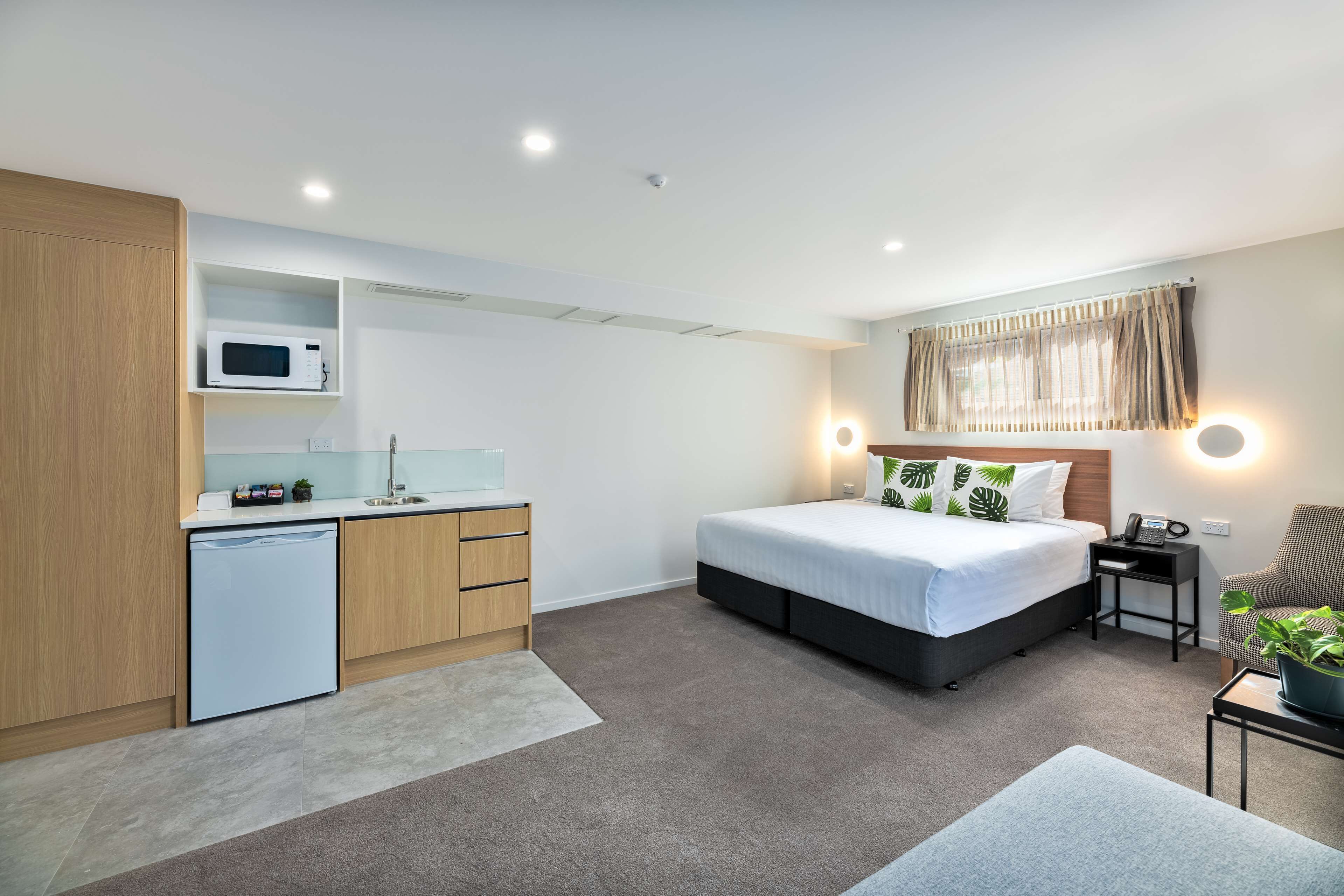 A compact hotel room with a kitchenette, a bed with a headboard, bedside tables, a chair, and a potted plant.
