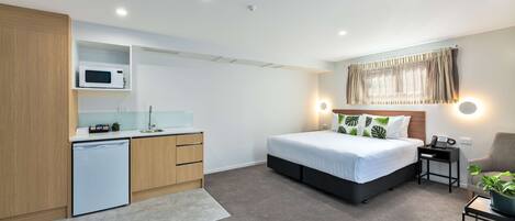 A compact hotel room with a kitchenette, a bed with a headboard, bedside tables, a chair, and a potted plant.