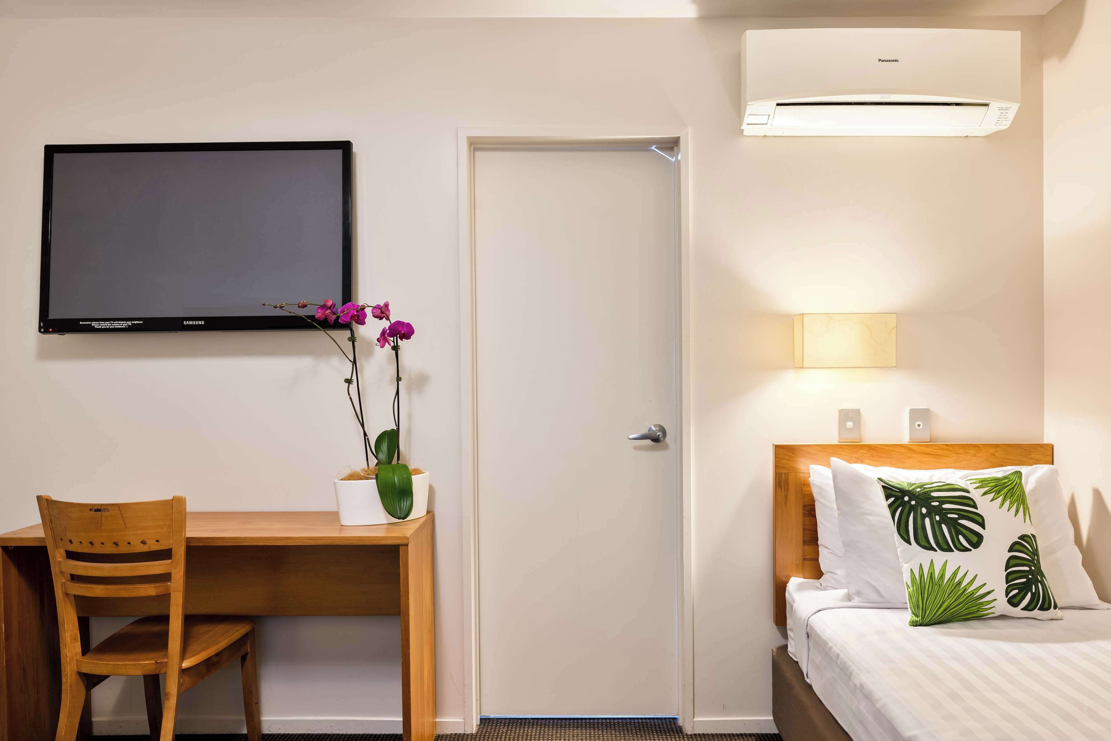 Habitación de hotel con un escritorio de madera, televisor, puerta, aire acondicionado y cama con almohadas con motivos tropicales.