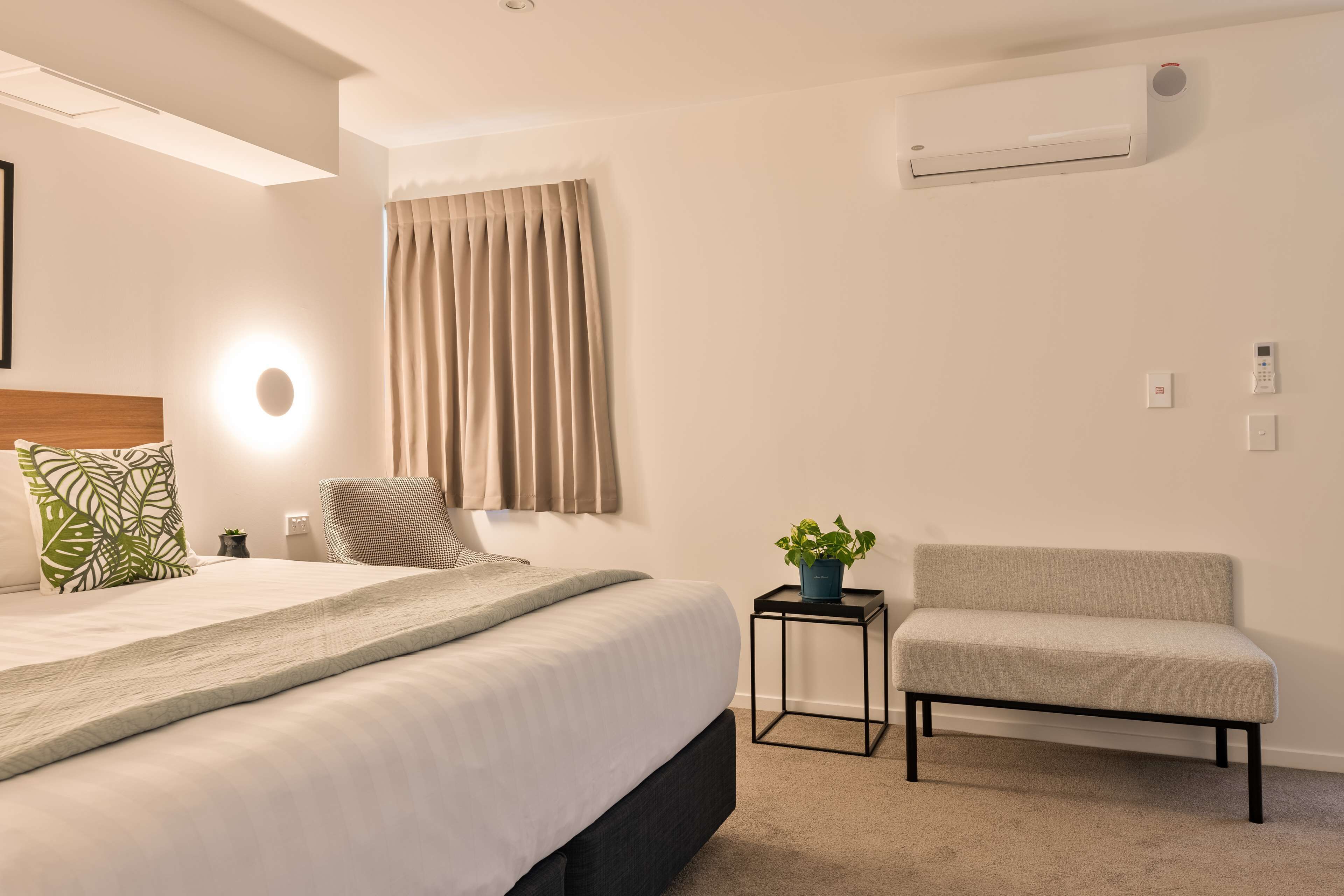 A modern bedroom with a bed, bedside table, chair, and an air conditioning unit.