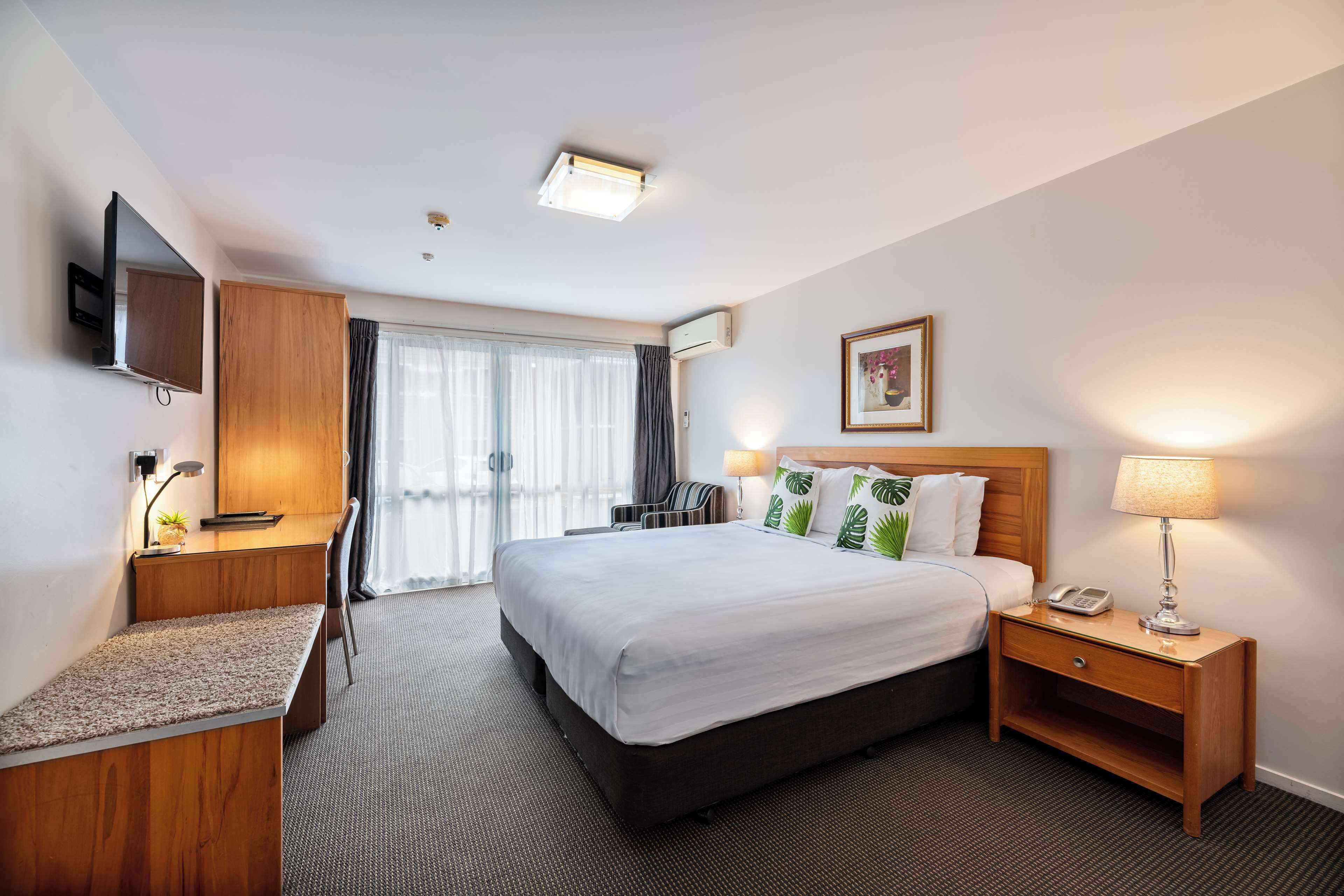 A hotel room with a bed, bedside table, lamp, desk, chair, and a TV mounted on the wall.