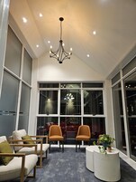 A modern interior with a high ceiling, recessed lighting, and a chandelier. There are two seating areas with sofas and armchairs, a small round table, and a potted plant. Large windows provide a view of the outside.