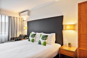 A hotel room with a large bed, a bedside table with a lamp, a chair, and a window with curtains.