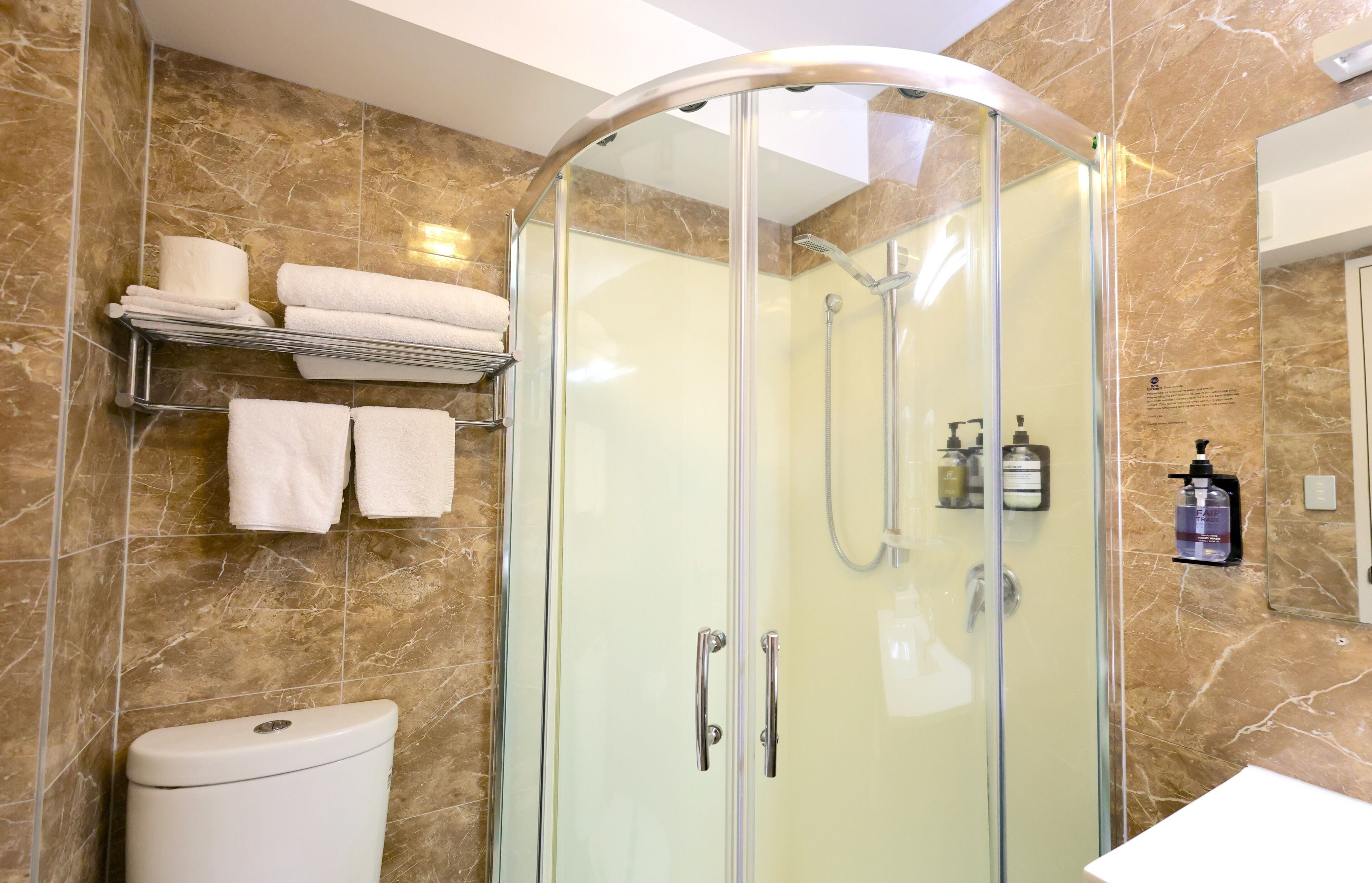 A modern bathroom with a glass-enclosed shower, marble walls, a toilet, and a shelf with towels.