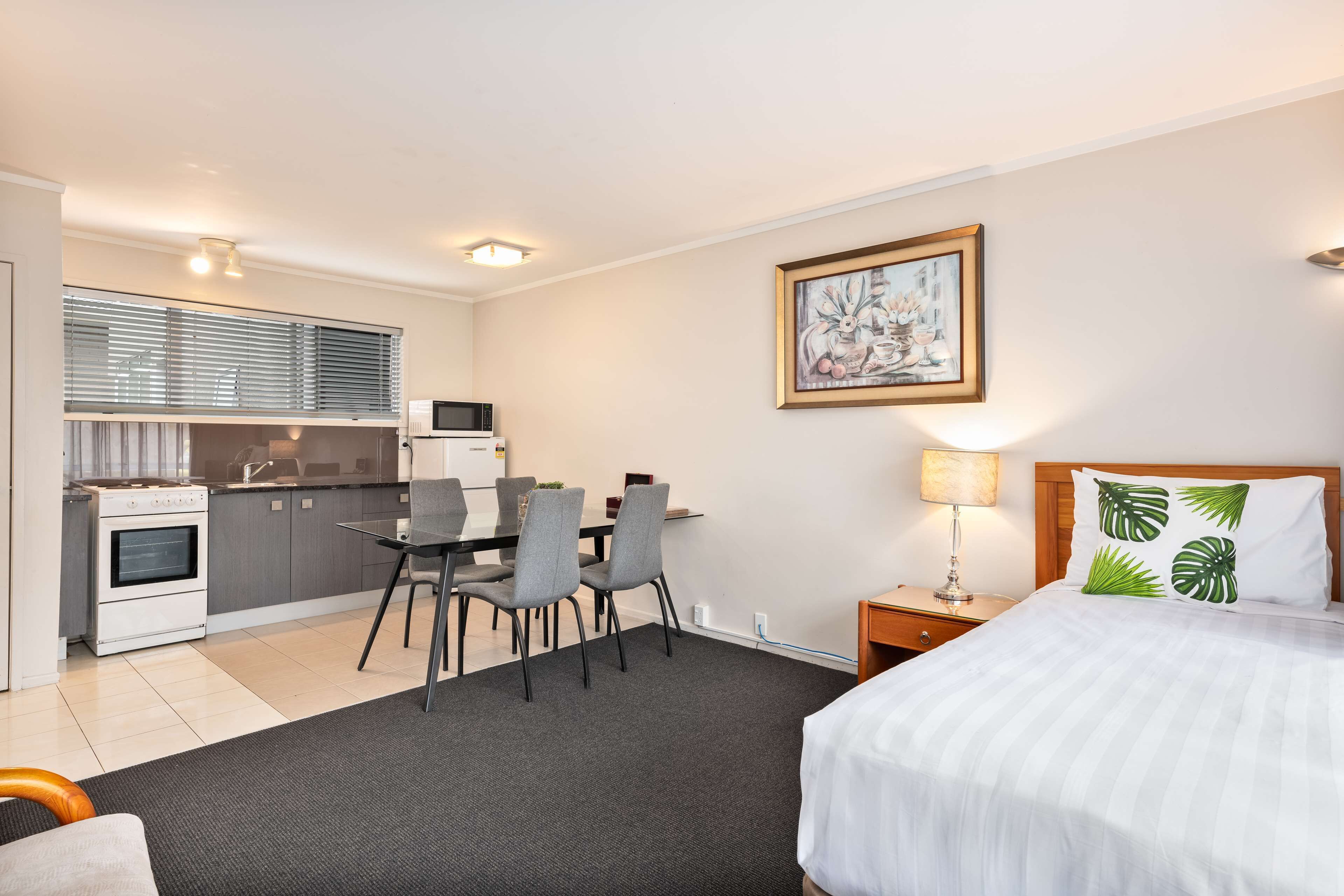 A hotel room with a bed, a desk with chairs, a kitchenette, and a painting on the wall.