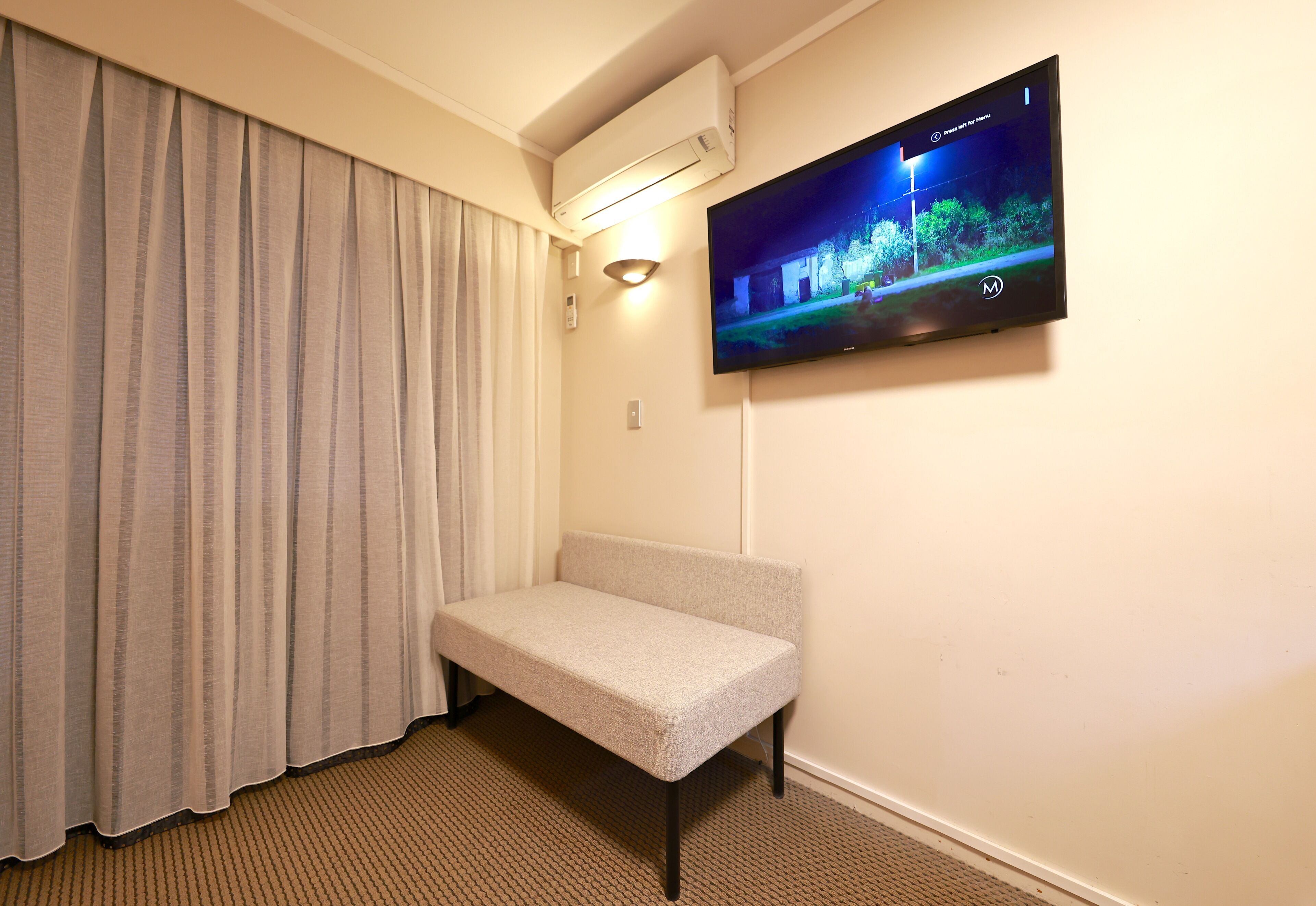 Een hotelkamer met een bed, een aan de muur bevestigde televisie, een raam met gordijnen en een airconditioner.