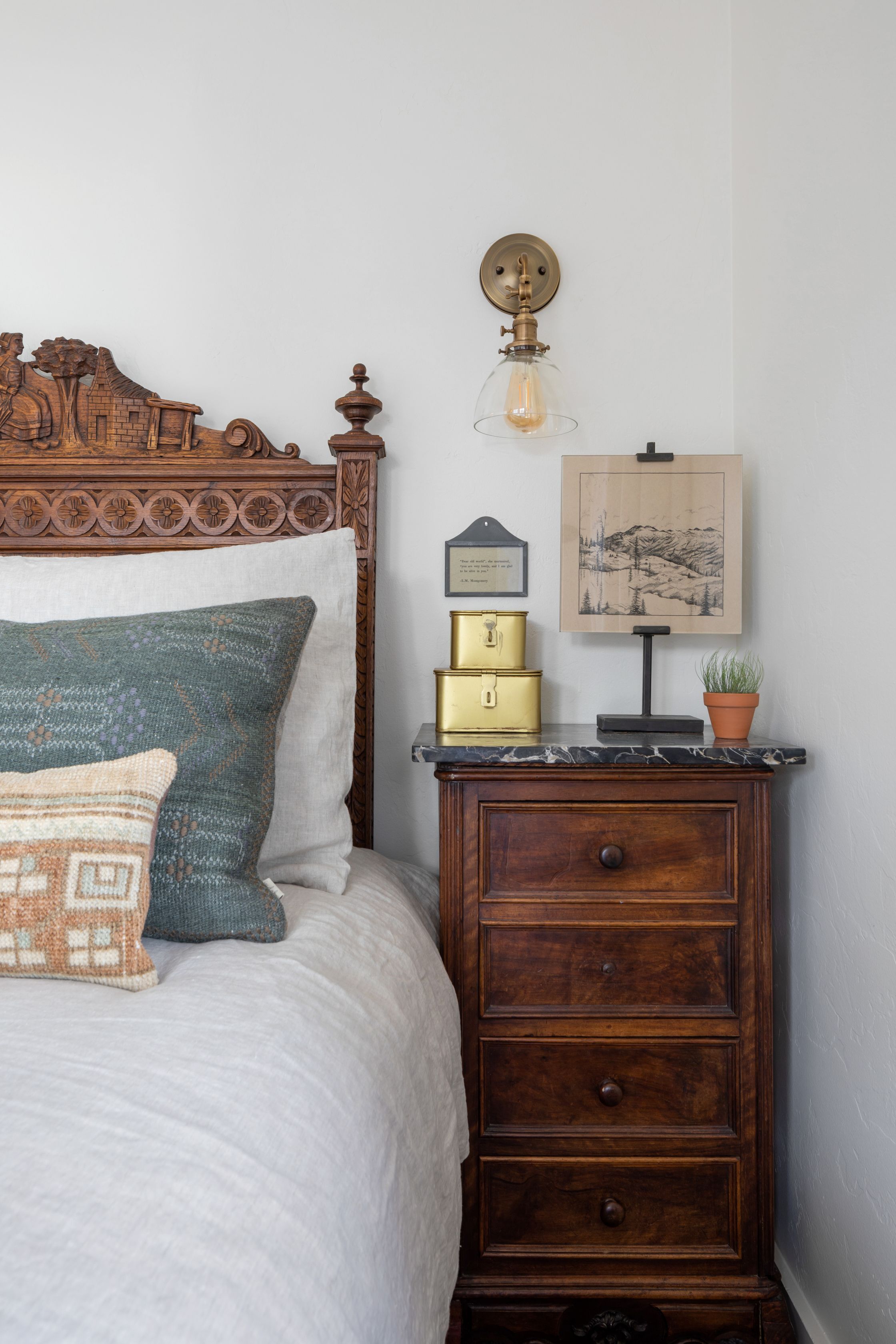 A neatly made bed with a wooden headboard, a bedside table with a lamp, a framed picture, and a small potted plant.