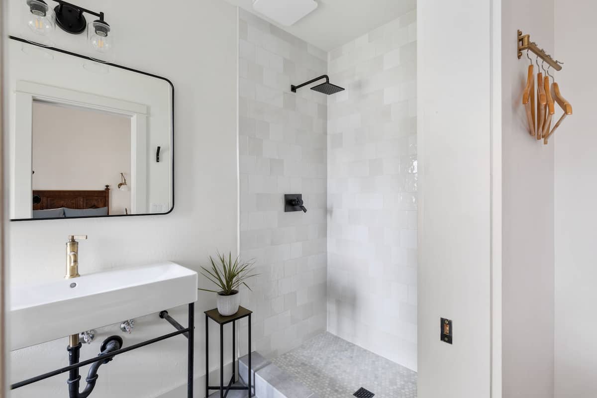 Une salle de bain moderne avec un lavabo blanc, un miroir, une douche avec un pommeau noir et une petite plante posée sur une table d’appoint.