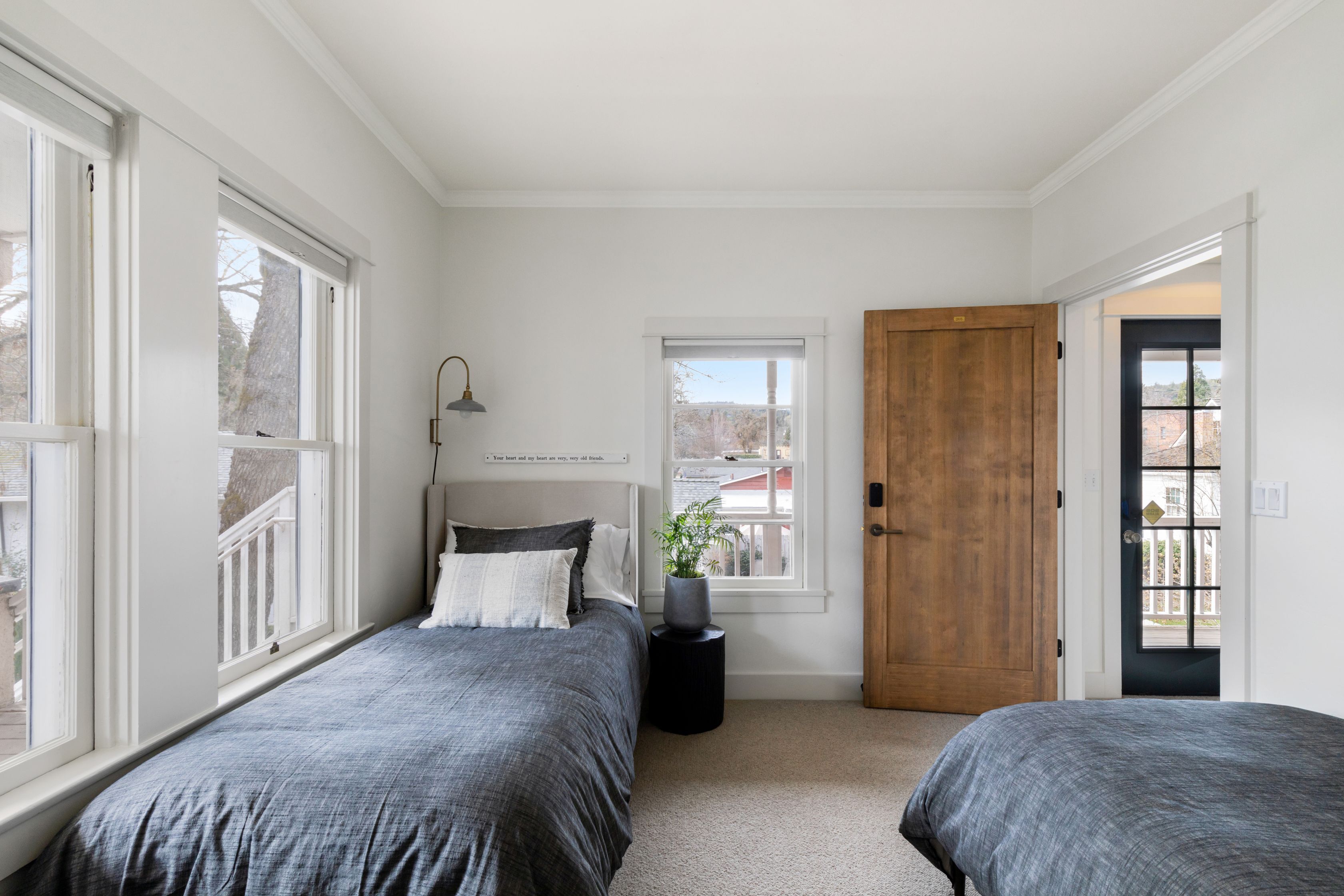 A bedroom with two beds, a window, a door, and a small table with a lamp.