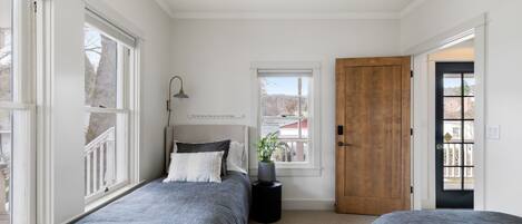 A bedroom with two beds, a window, a door, and a small table with a lamp.