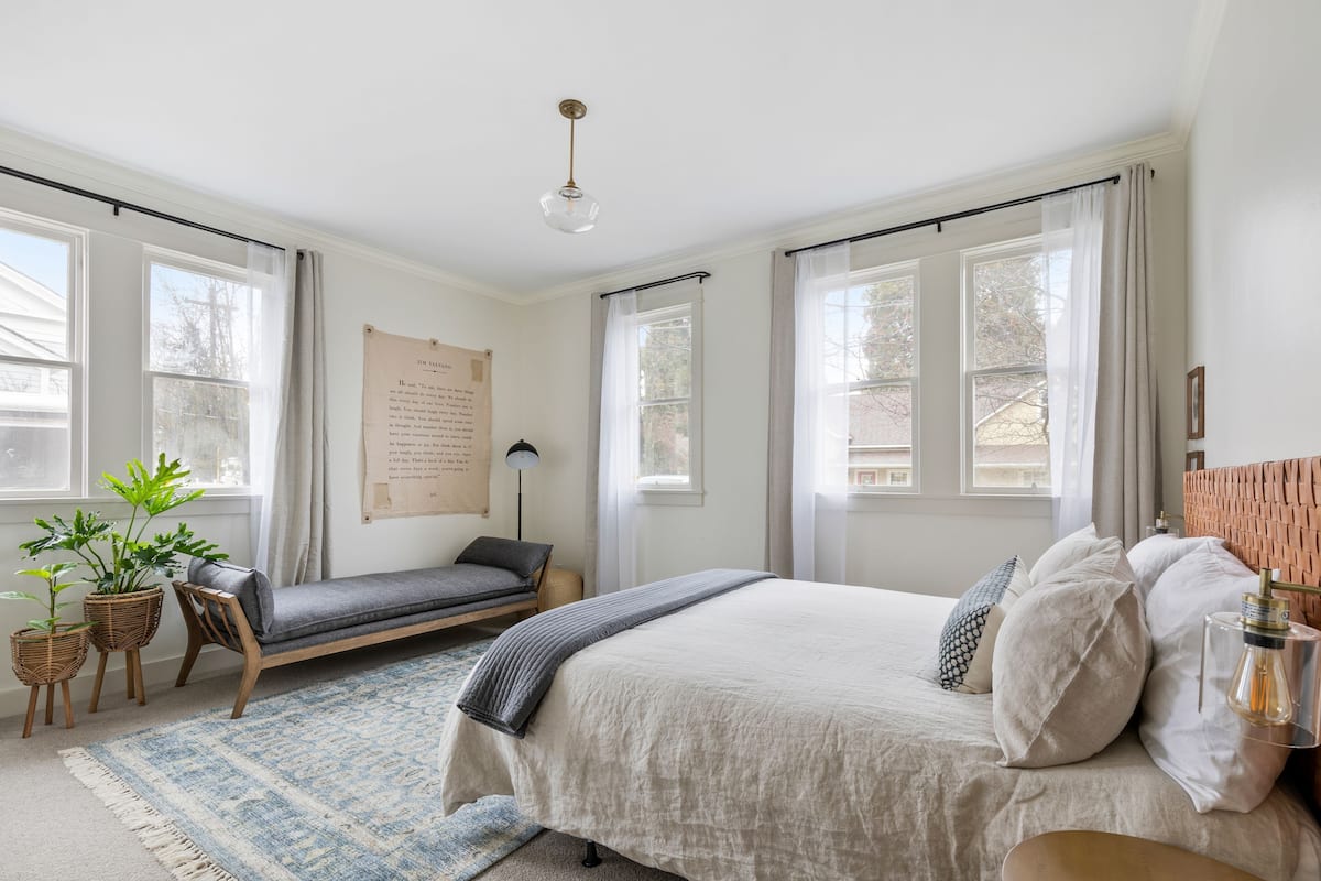 Une chambre à coucher comprenant un lit, un banc, deux plantes en pot, une lampe et un mur sur lequel est affiché un avis.