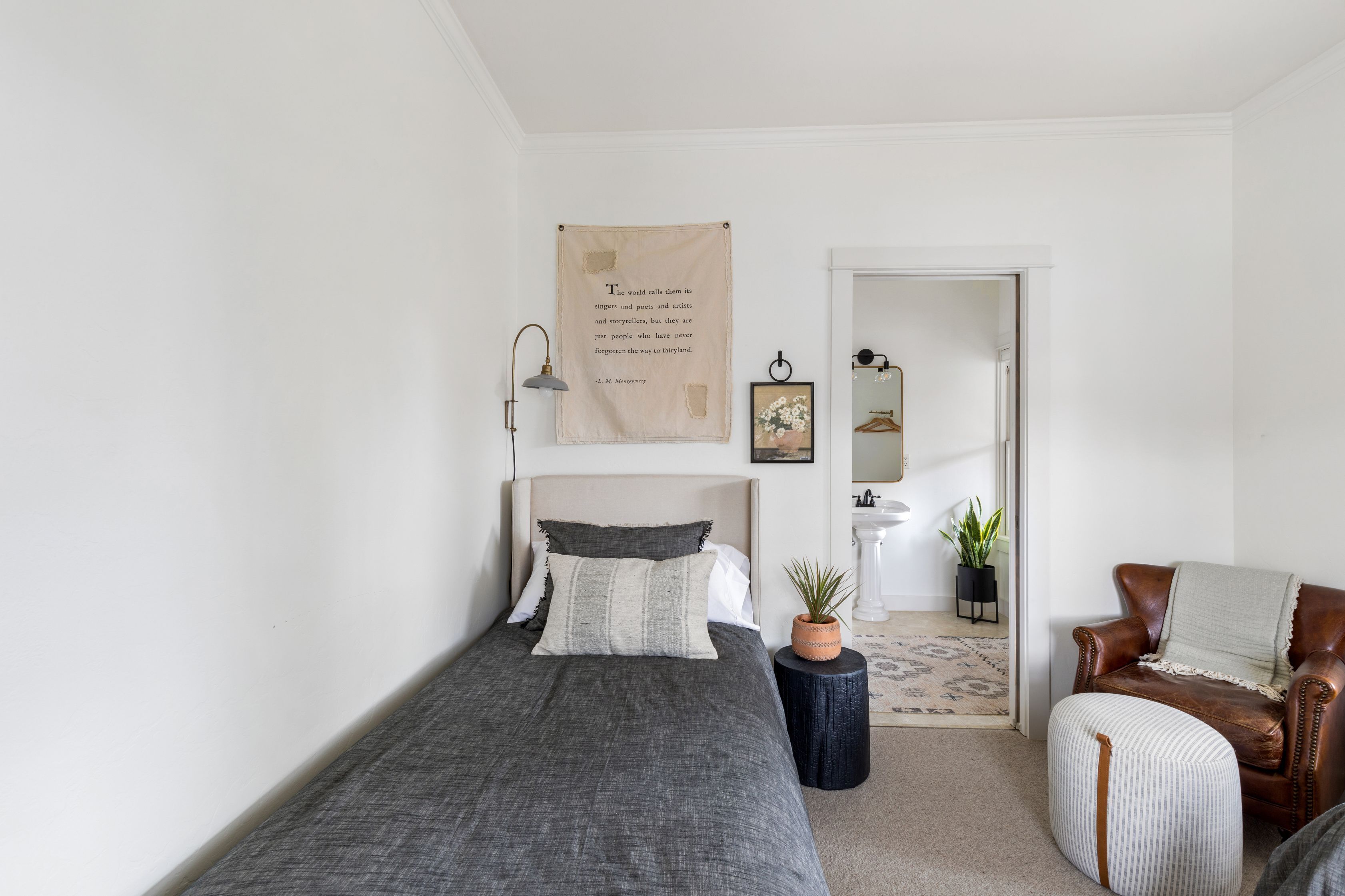 Une chambre à coucher comprenant un lit, une table de chevet, une plante en pot, une chaise et une porte donnant sur une autre pièce.