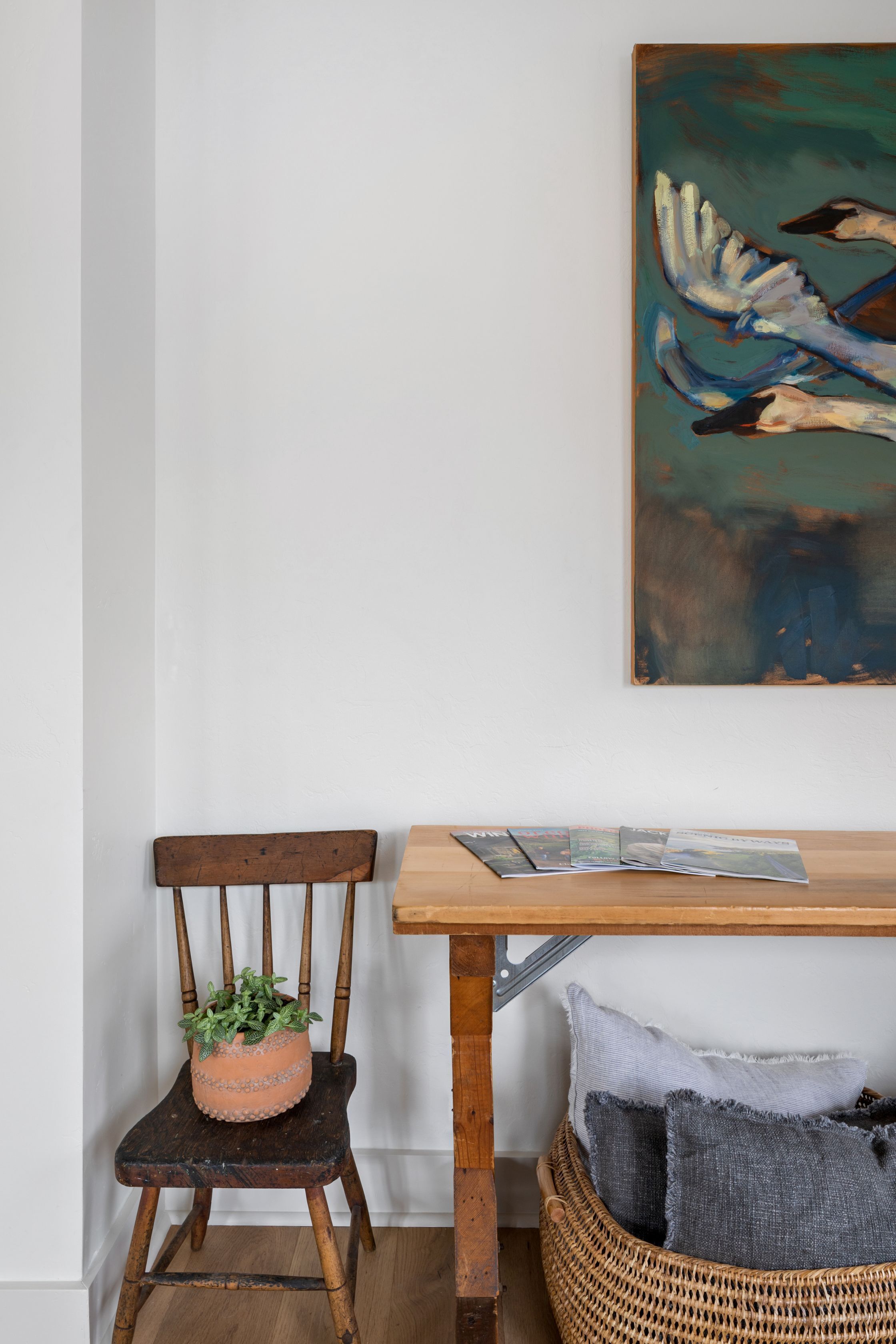 A wooden chair with a potted plant, a wicker basket with pillows, and a painting on the wall.