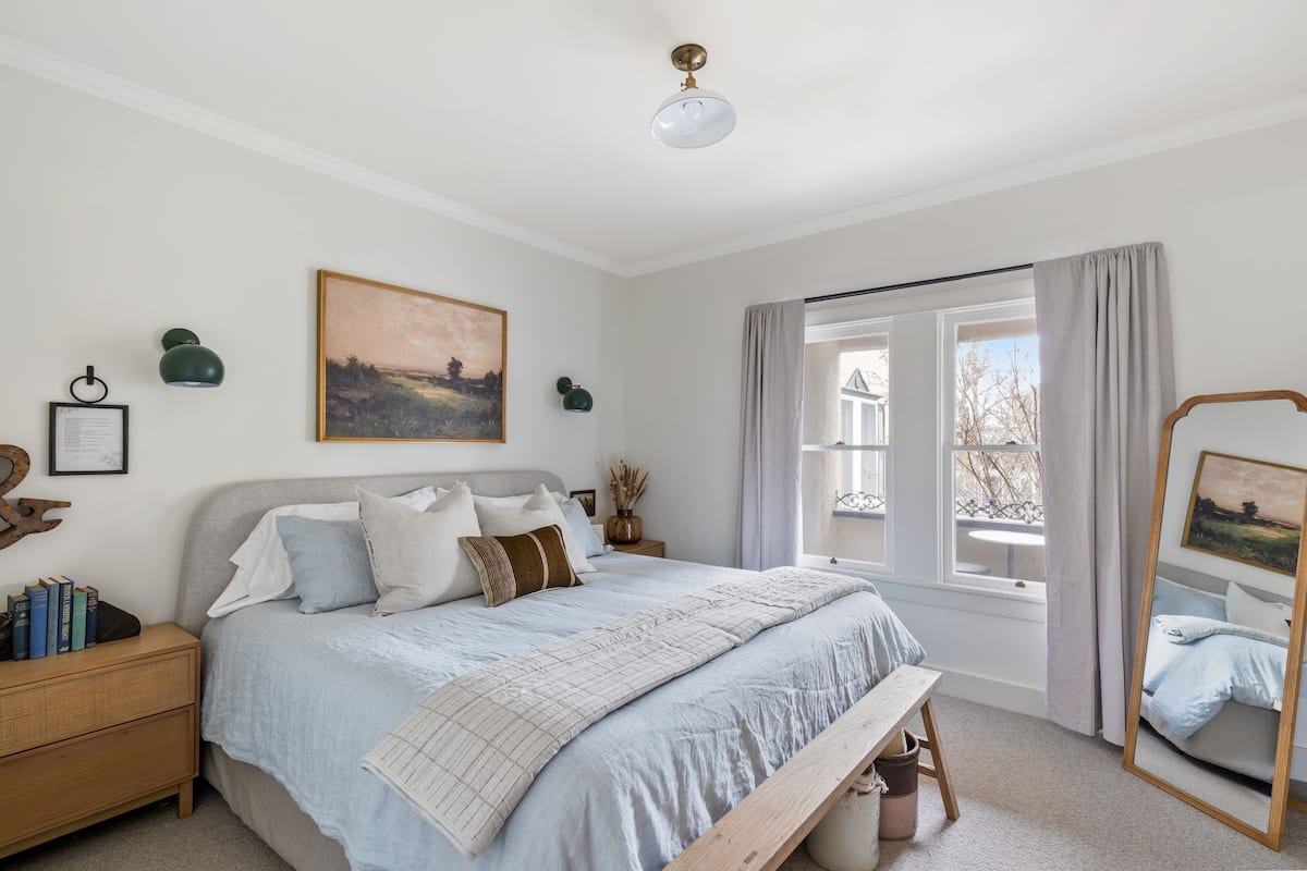 Une chambre à coucher avec un lit, des tables de chevet, une fenêtre avec des rideaux, un luminaire au plafond et un tableau encadré au mur.