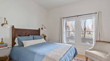 A bedroom with a large bed, a chair, a window with curtains, and a view of the outside.
