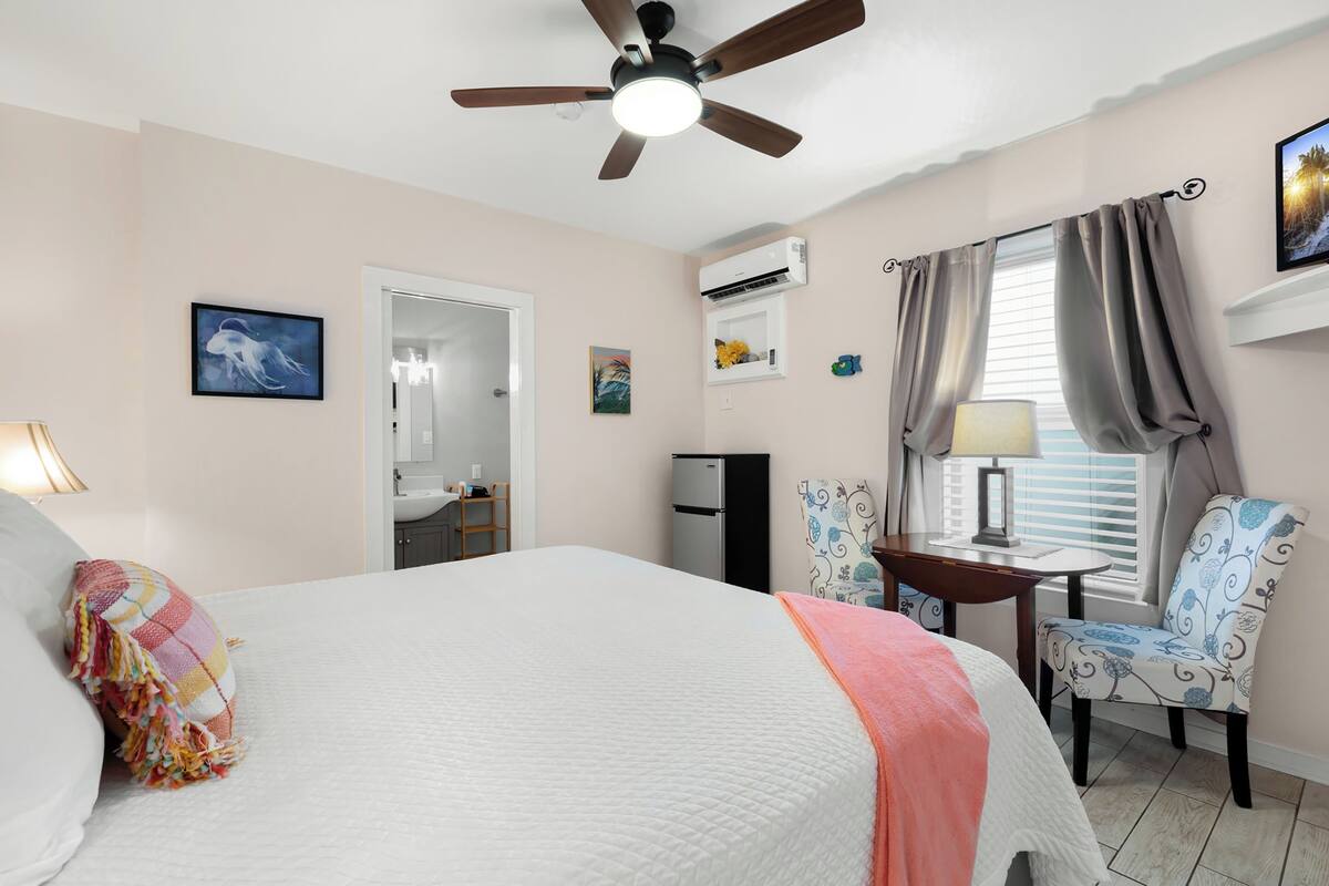 A bedroom with a bed, a ceiling fan, a desk with a chair, an air conditioning unit, and a window with curtains.