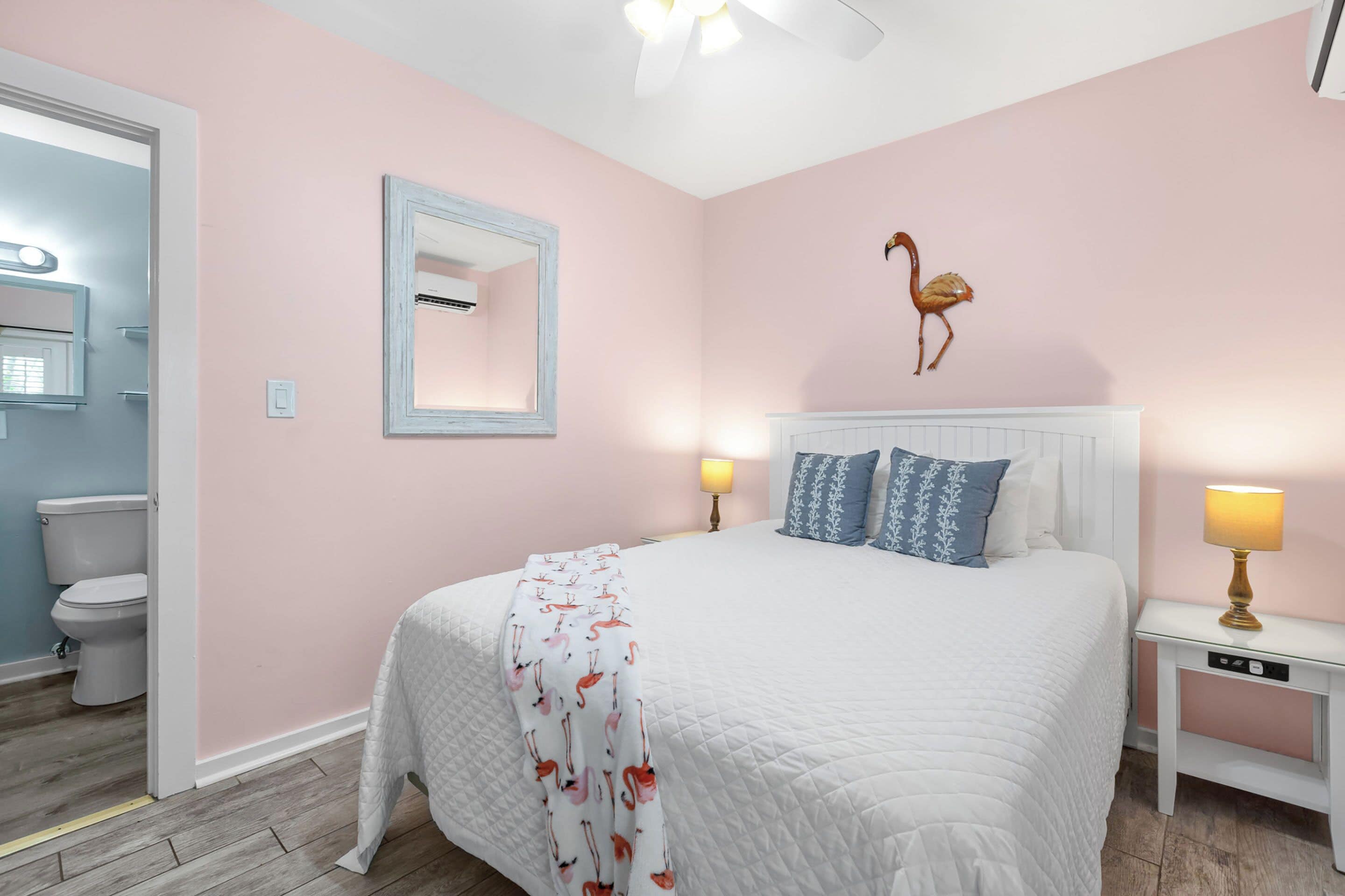 A bedroom with a white bed, a nightstand with a lamp, a wall-mounted mirror, and a wall decoration of a flamingo.