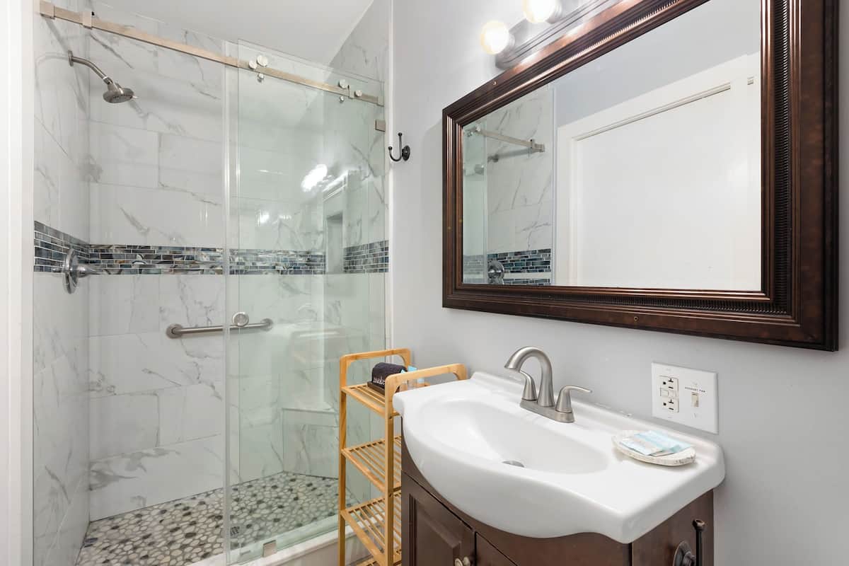 A bathroom with a glass-enclosed shower, a white sink with a mirror, and a wooden shelf.