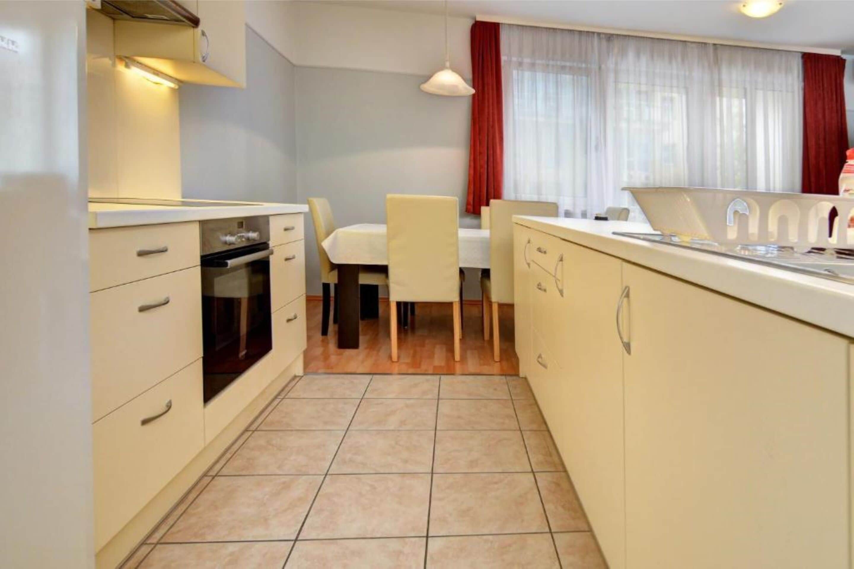 Una cocina compacta con horno integrado, un área de comedor con mesa y sillas, y un ventanal con cortinas.
