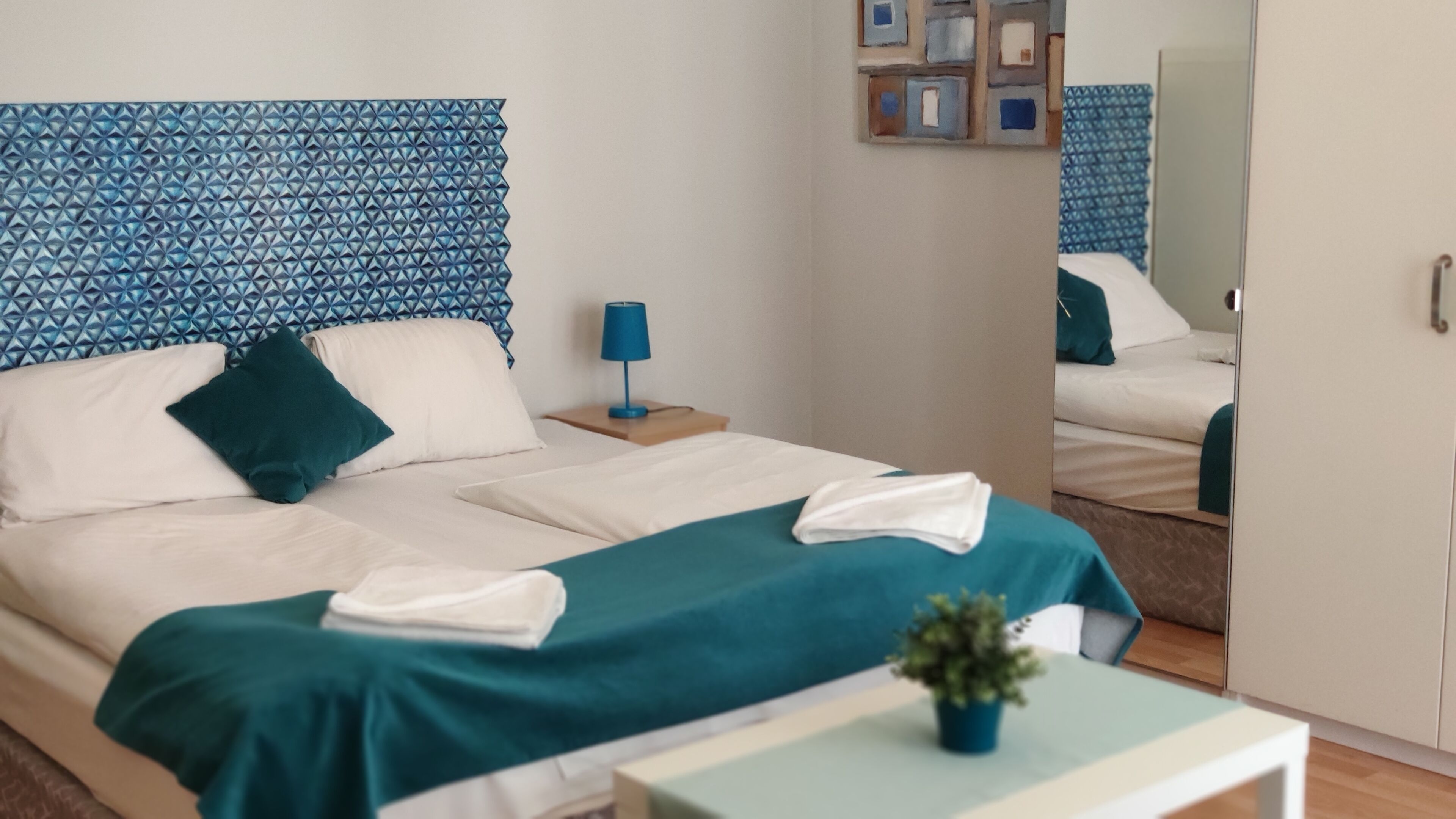 Una cama bien hecha con colcha color turquesa y sábanas blancas, una mesita de noche con lámpara, una planta en maceta sobre una mesita auxiliar y un espejo que refleja parte de la habitación.