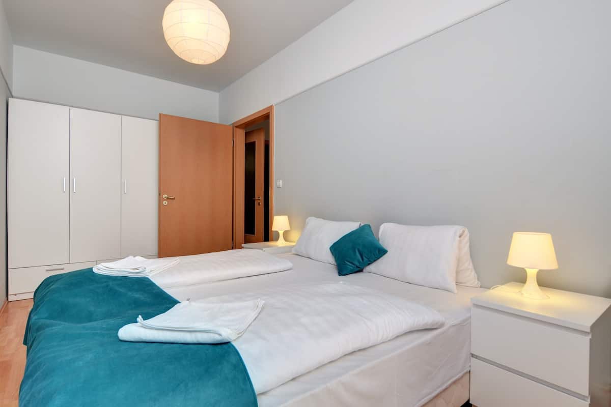 A bedroom with two beds, a wardrobe, a nightstand with a lamp, and a door leading to another room.
