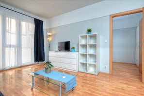 Un salon moderne avec une table basse en verre, un téléviseur à écran plat, une étagère blanche et une porte donnant sur une autre pièce.
