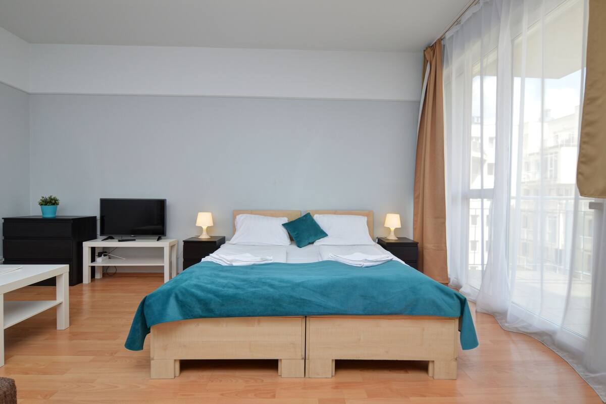 A neatly arranged bedroom with a bed, bedside tables, a TV, and a window with curtains.