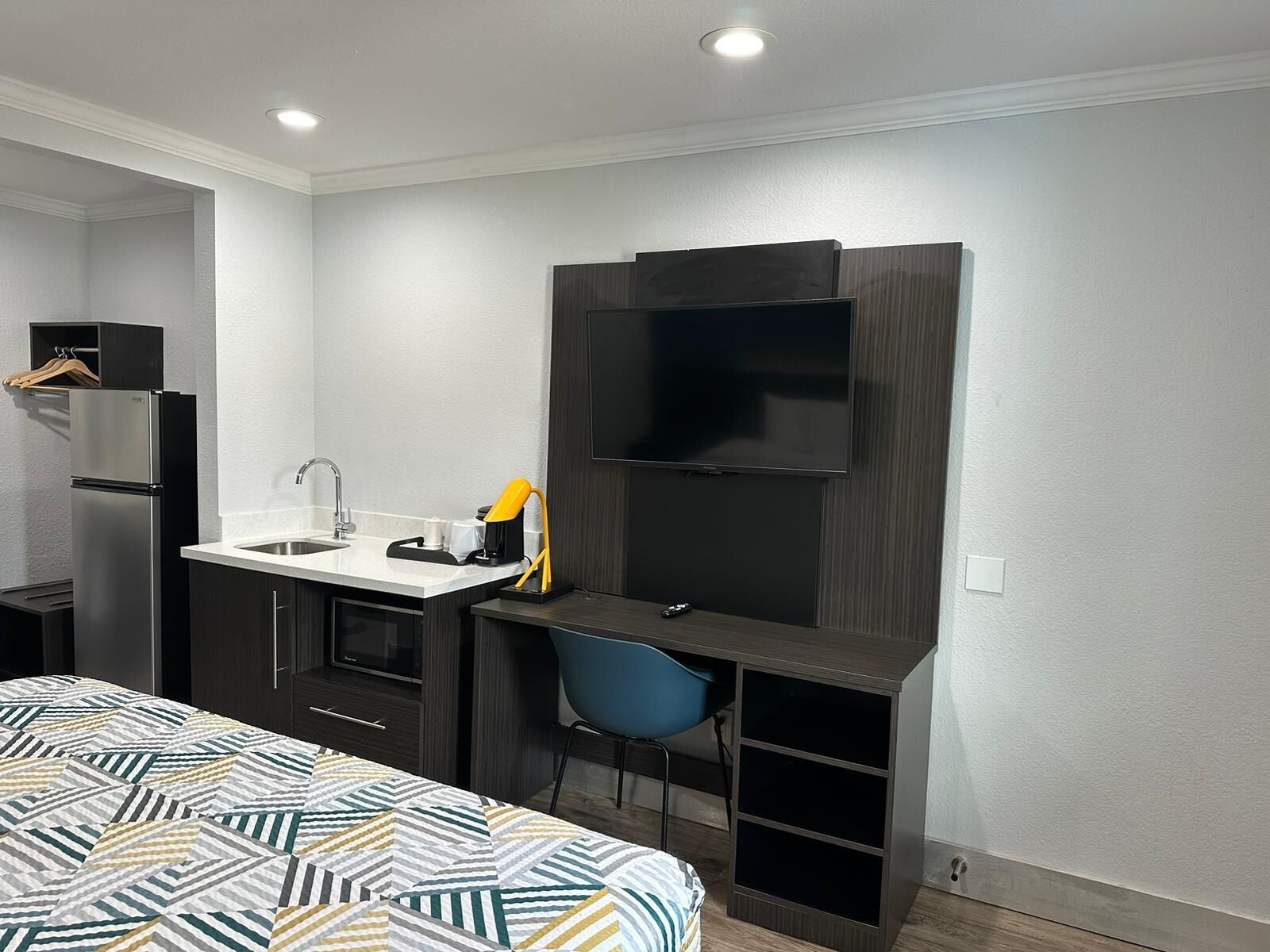 Een kleine hotelkamer met kitchenette, een bed met een beddegoed met patroon, een bureau met stoel, een flatscreen televisie en een inbouwplank.