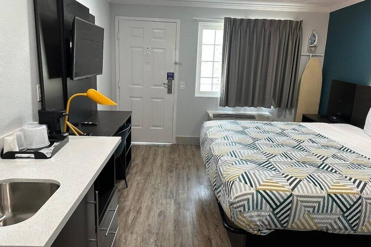 A hotel room with a bed, a desk with a chair, a TV mounted on the wall, a window with curtains, and a sink.