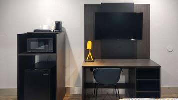 Una habitación funcional con escritorio, microondas, cafetera, silla y televisor montado en la pared.