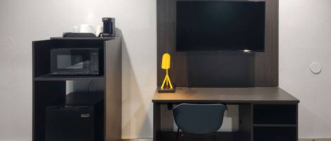 A compact room with a desk, a microwave, a coffee maker, a chair, and a TV mounted on the wall.